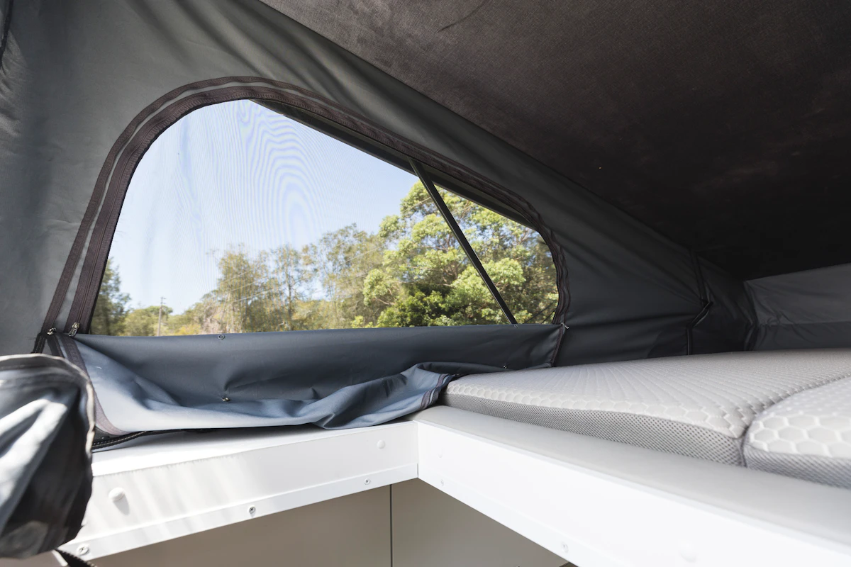 Interior view of a campervan roof tent with mattress and mesh window overlooking trees outside.