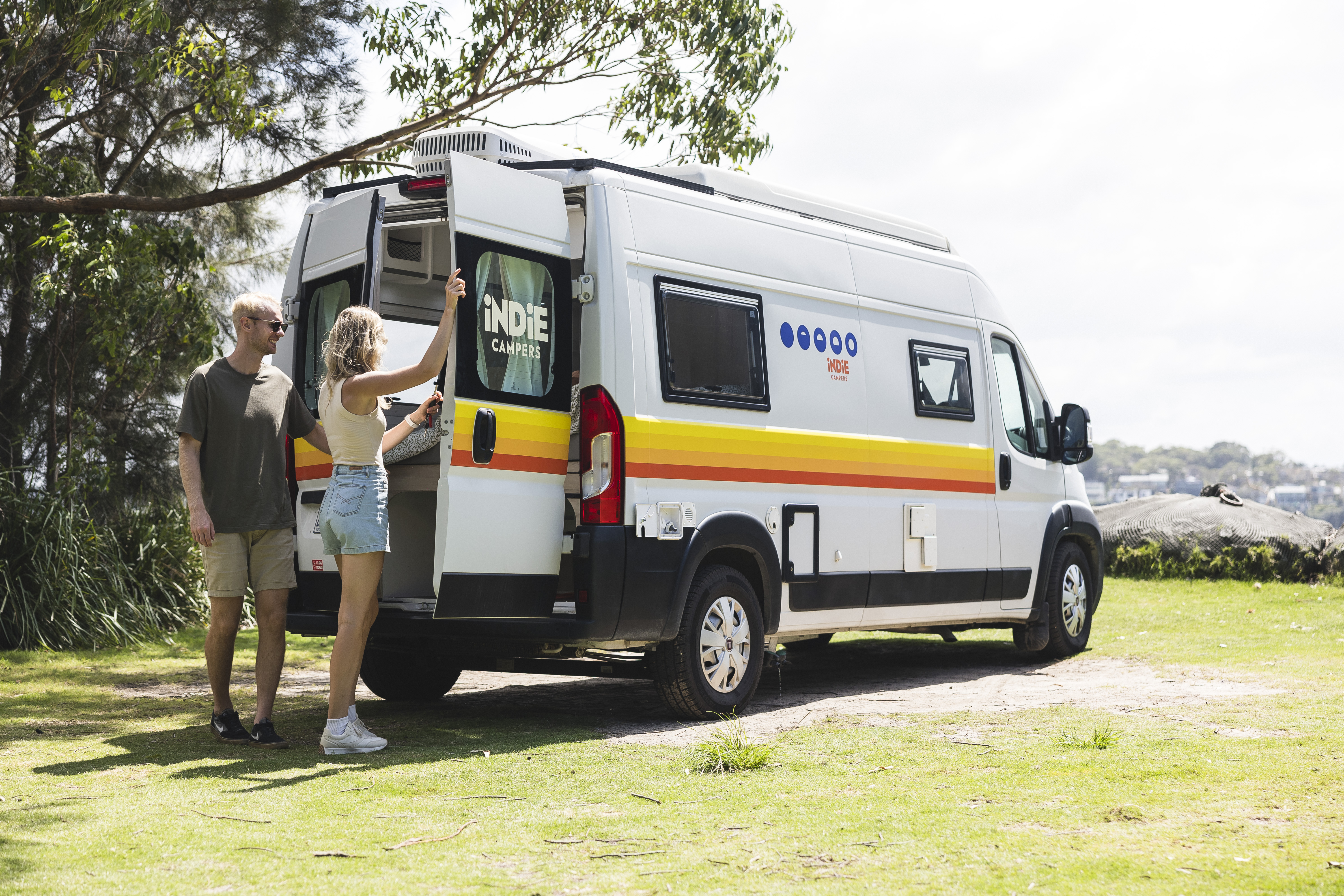 Two people standing outside a parked Indie Campers van in a grassy, outdoor setting with trees and hills.