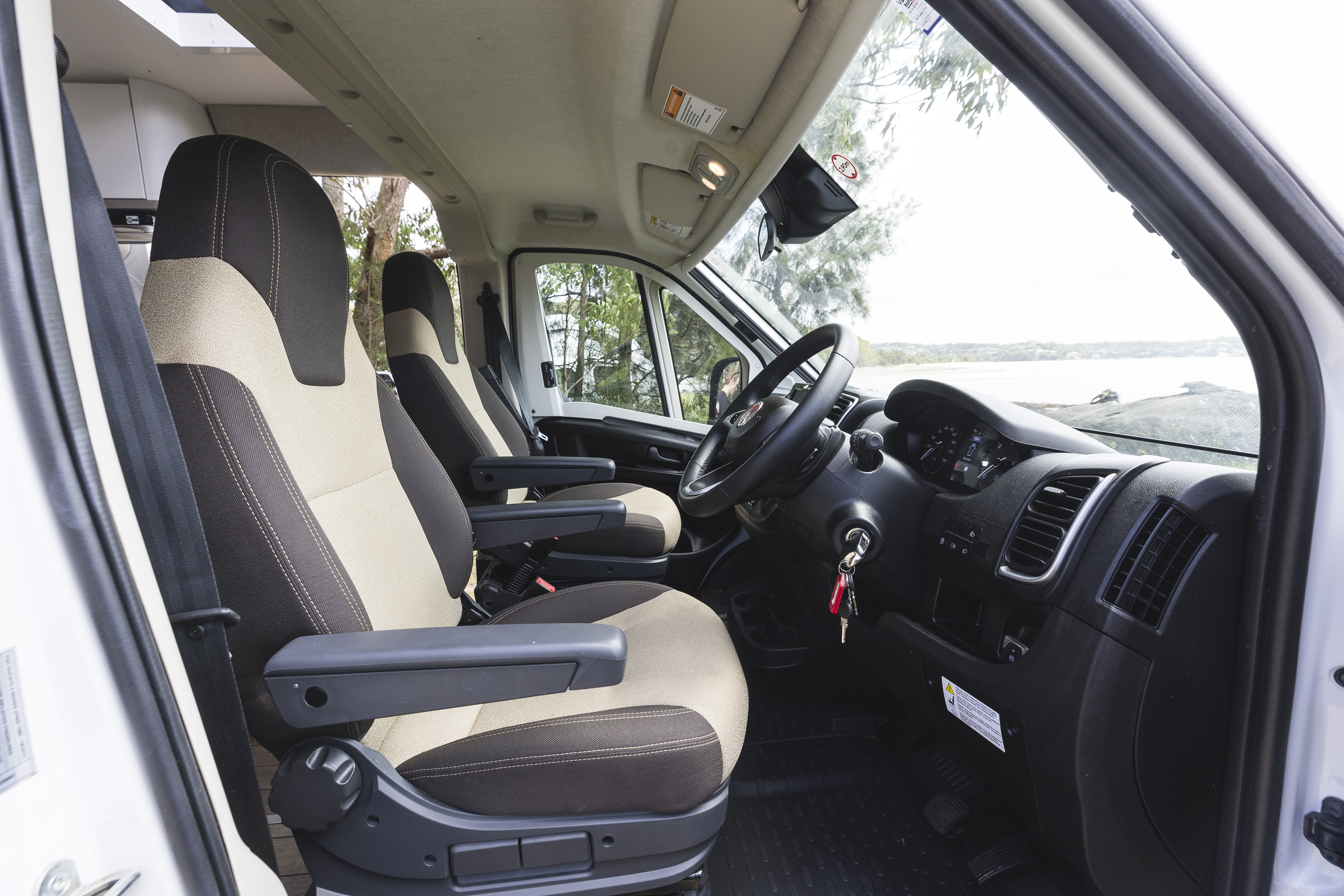 Interior view of a campervan driver’s cabin with two seats, steering wheel, and dashboard, parked near water.