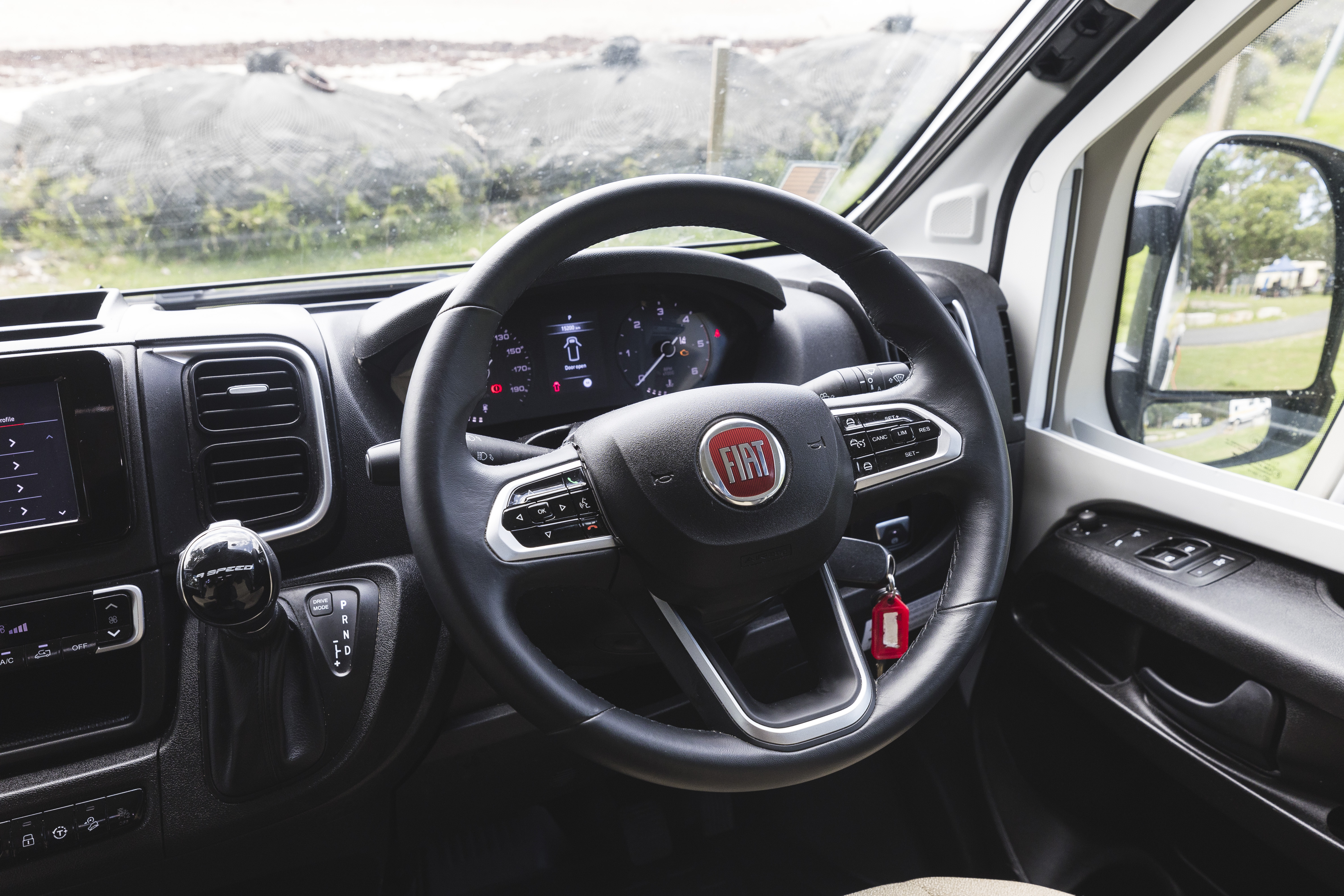 Fiat campervan interior showing steering wheel, dashboard, gear shift, and driver’s side window with scenic outdoor view.