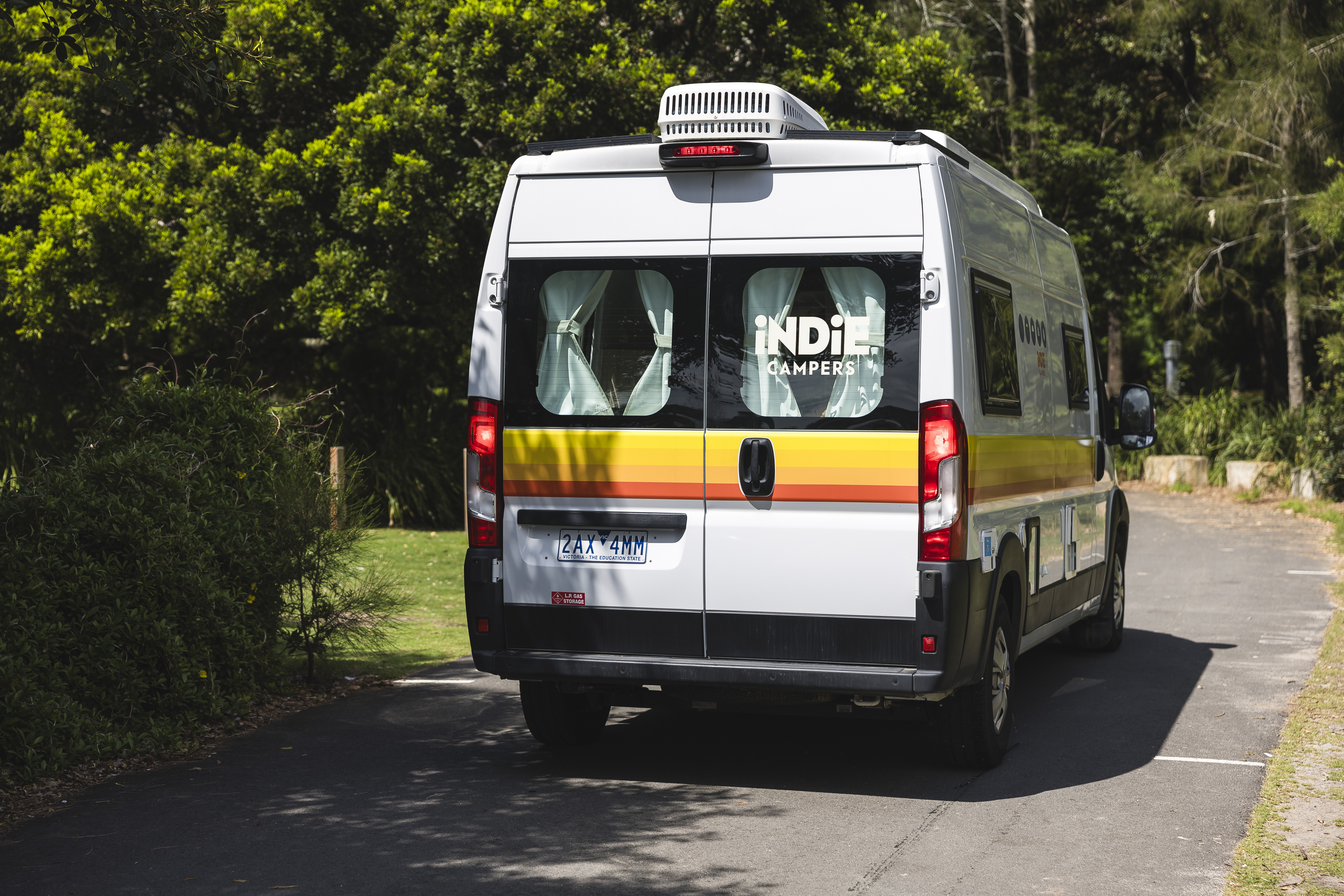 White Indie Campers van parked on a paved road surrounded by trees and greenery on a sunny day.