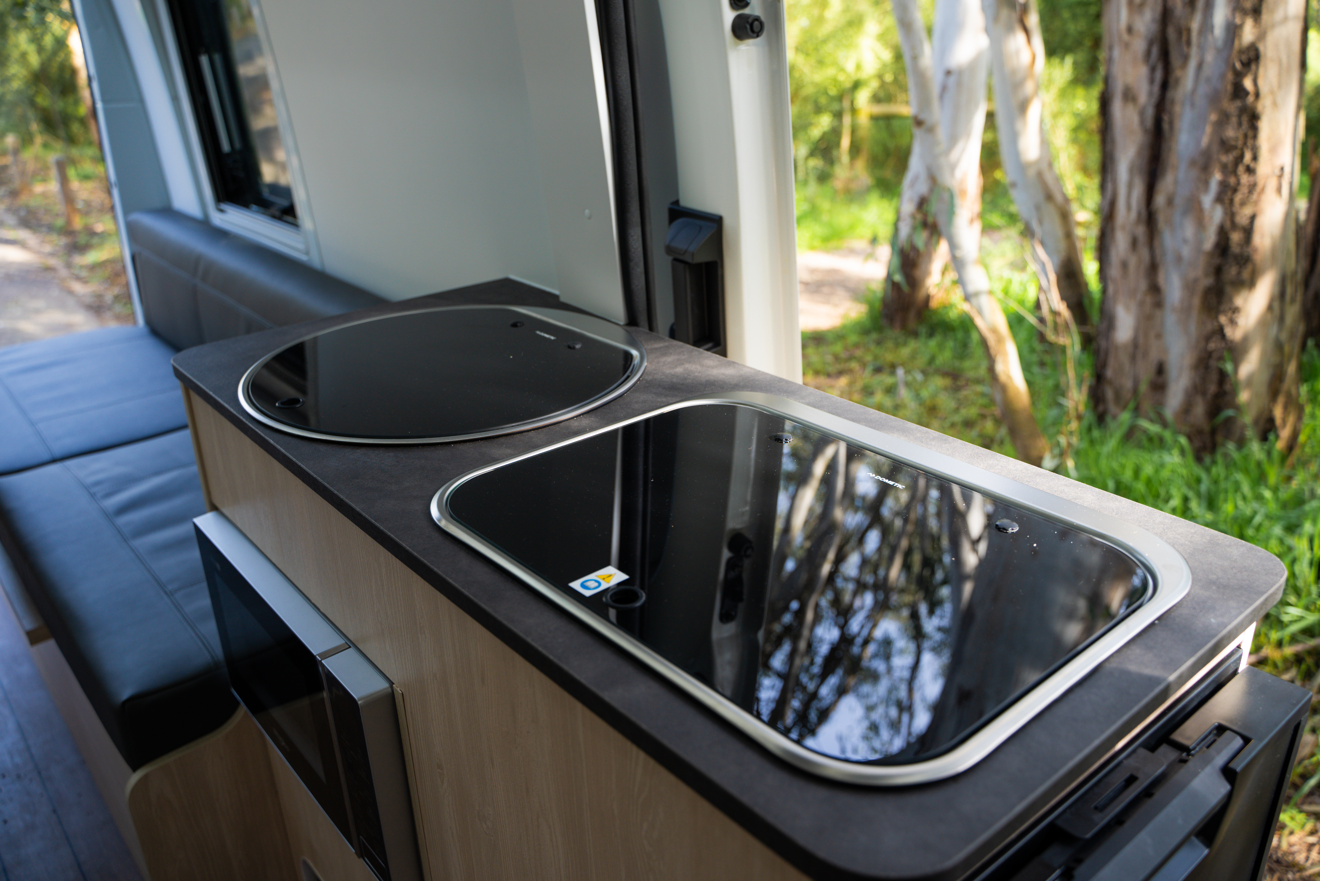 Modern campervan kitchenette with glass-covered stove and sink, black countertop, and adjacent seating area, surrounded by nature.