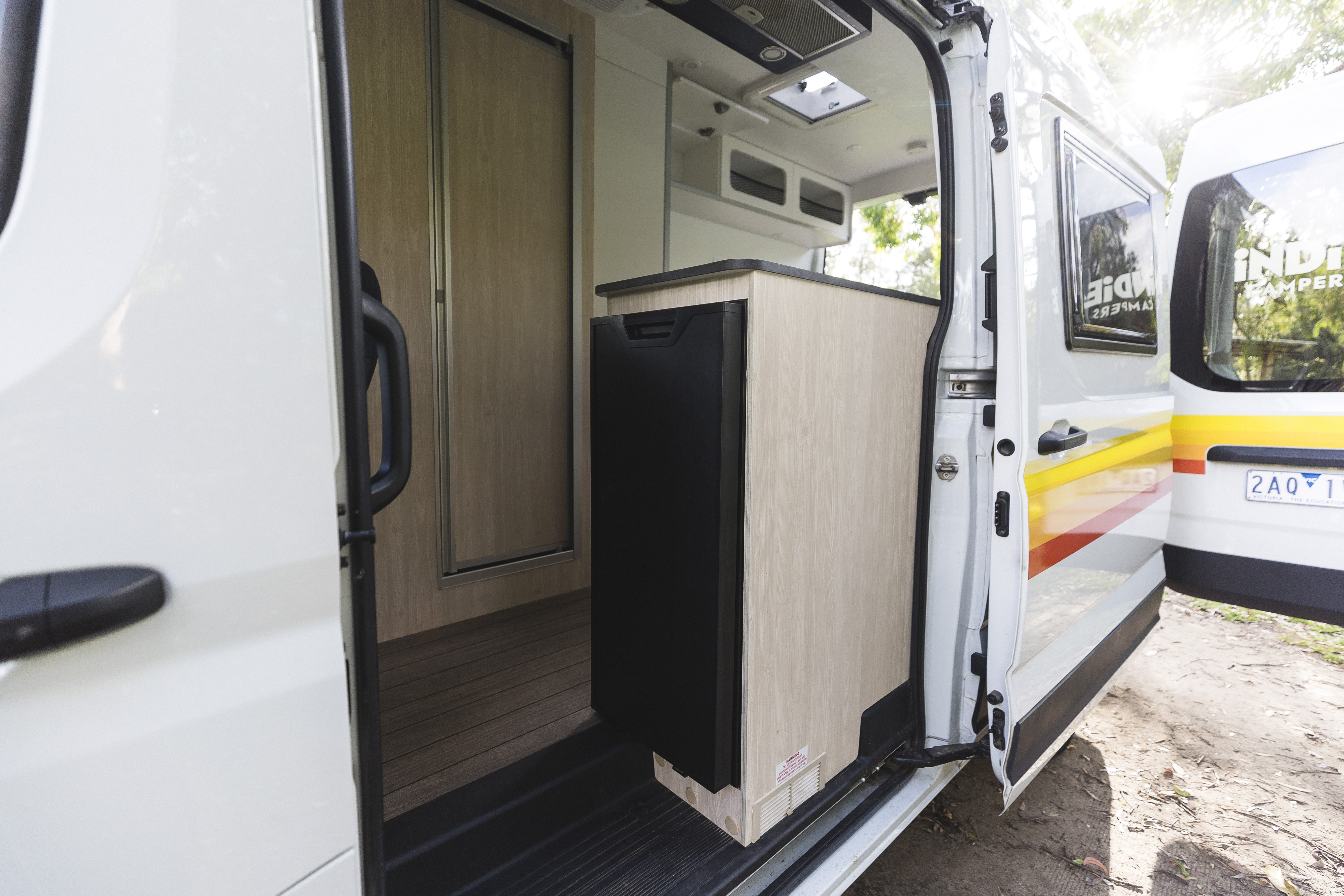Campervan open side door showing compact kitchenette with wooden cabinet, refrigerator, and interior details.