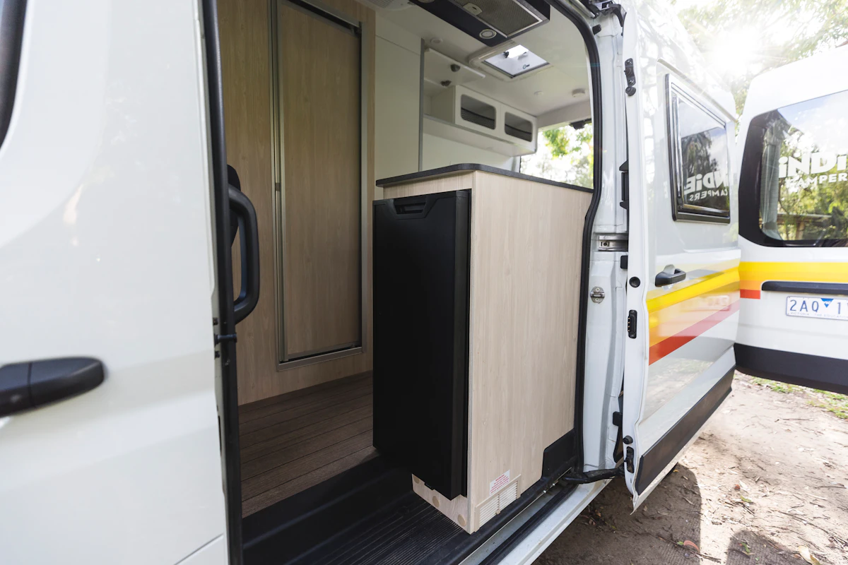 Campervan open side door showing compact kitchenette with wooden cabinet, refrigerator, and interior details.