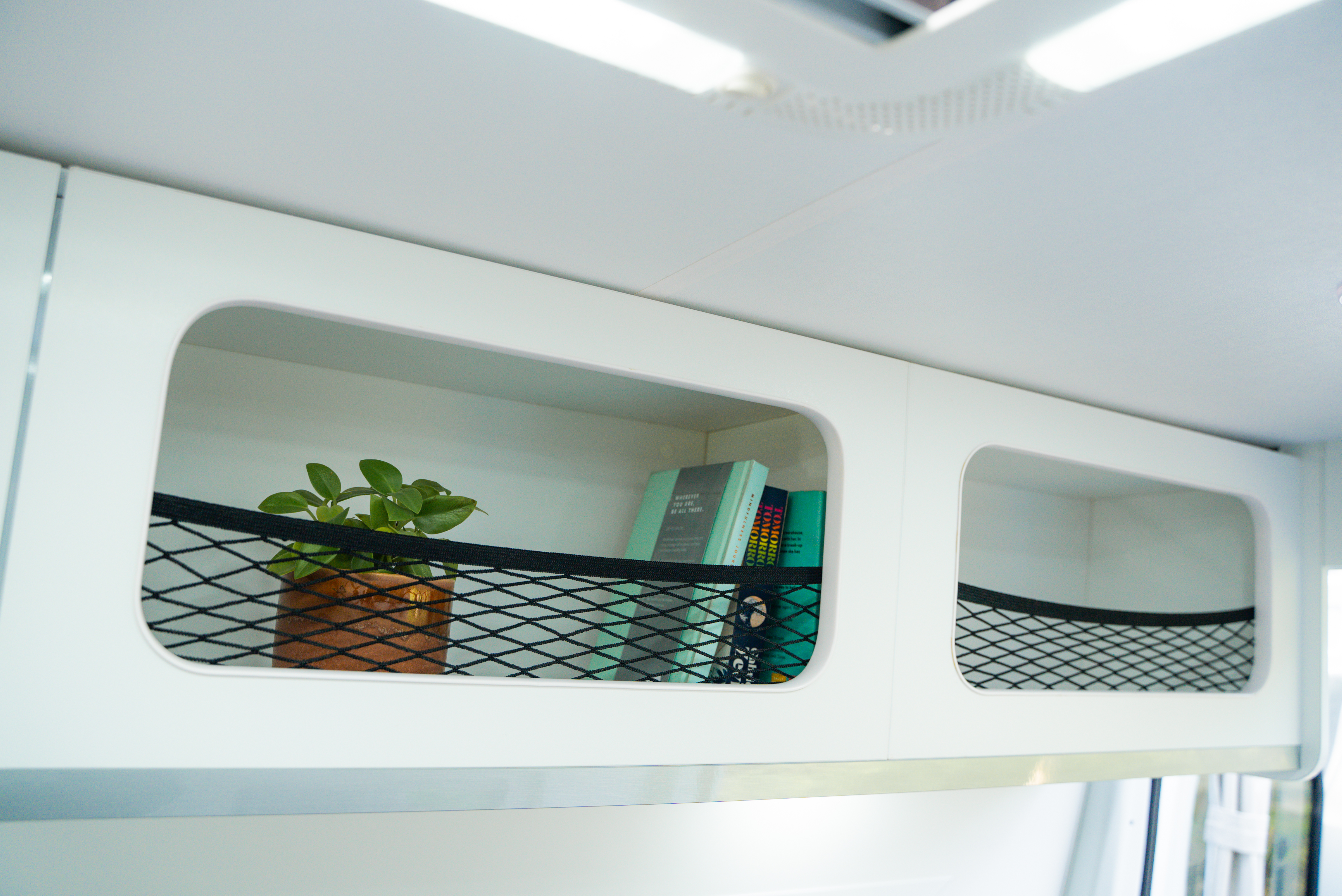 White campervan storage shelf with a small potted plant and books, secured by a black netting.