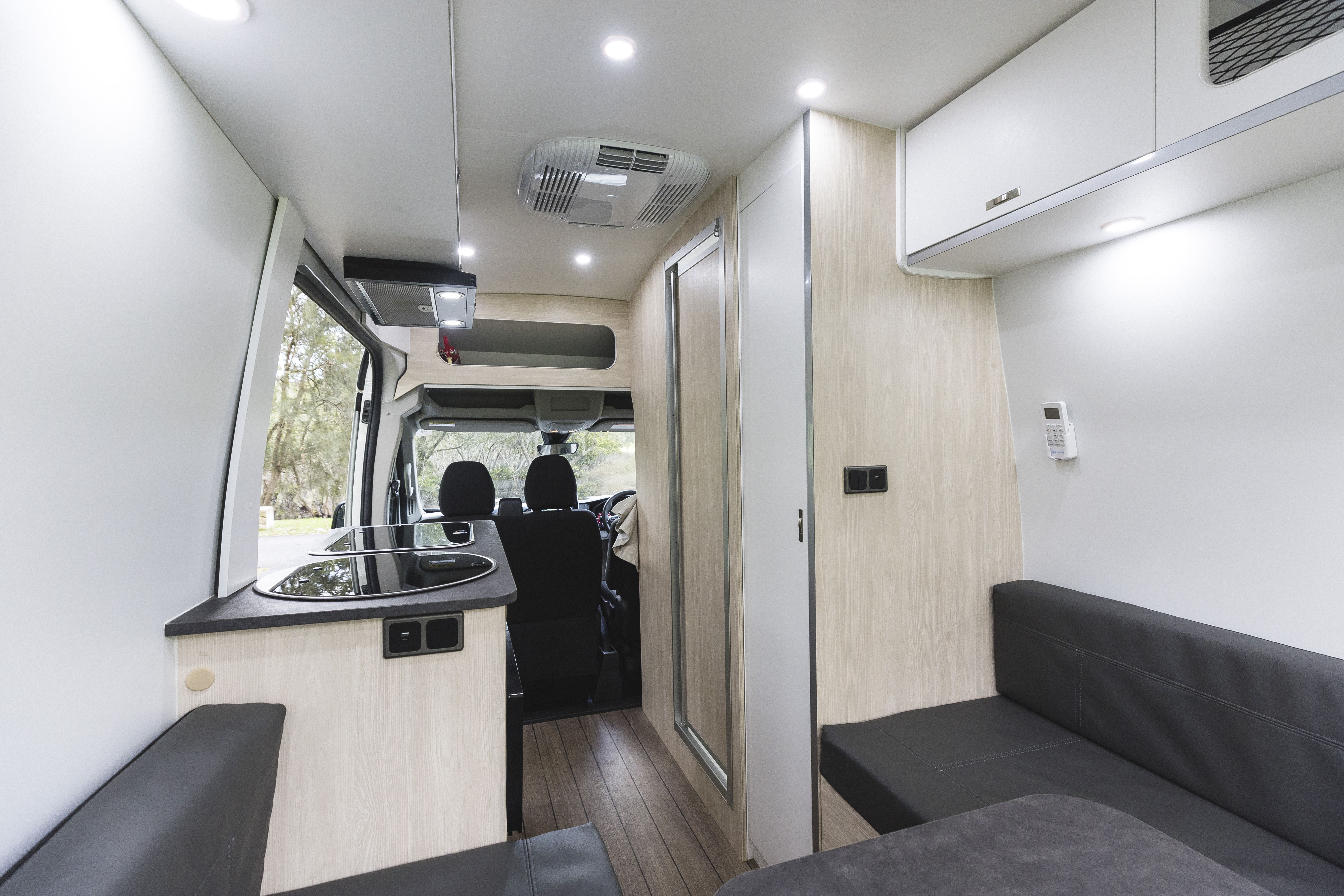 Modern campervan interior with kitchenette, seating area, cabinets, wood flooring, and front driver’s cabin visible through open layout.