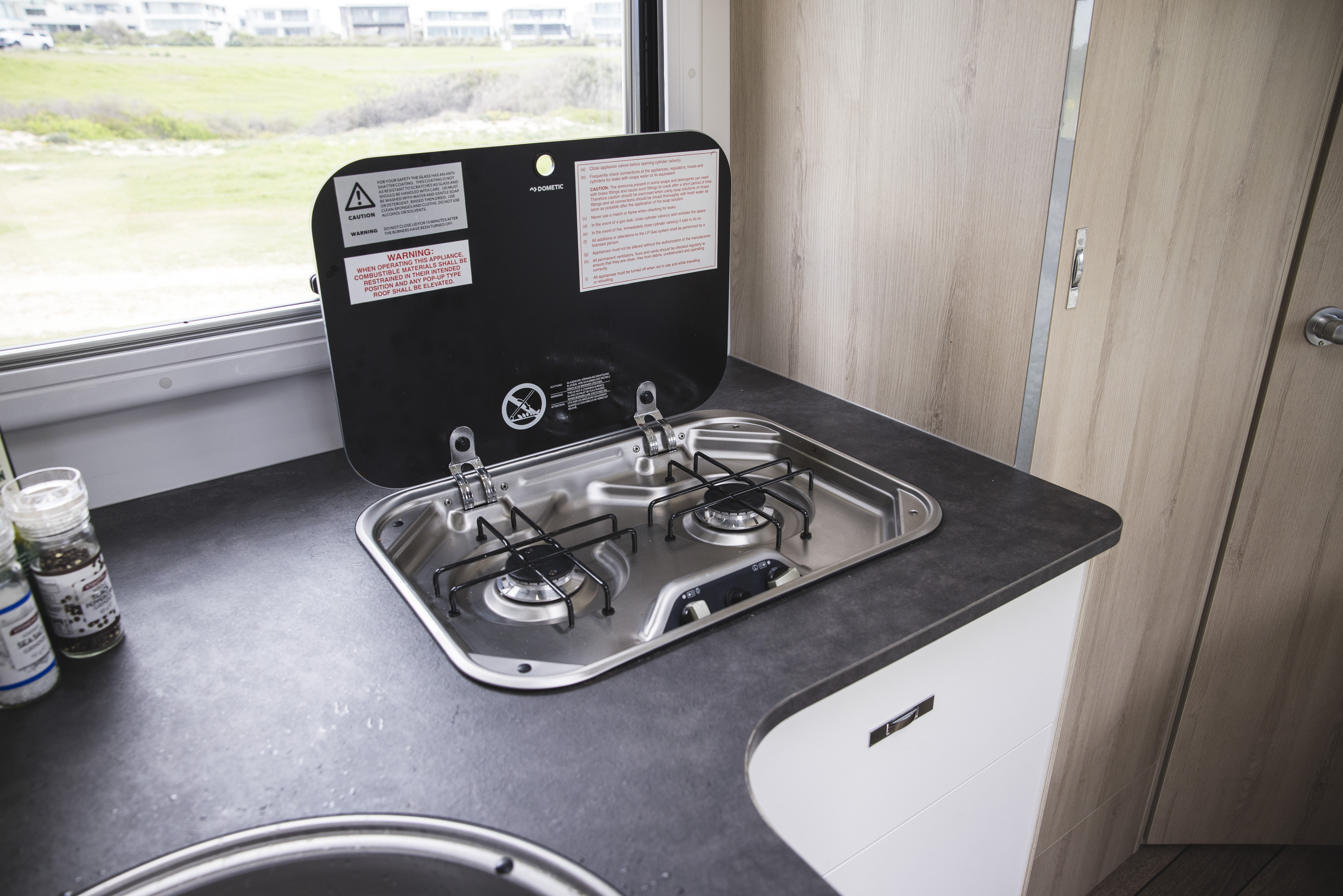 Two-burner gas stove with glass cover inside a campervan kitchen, next to window and countertop.