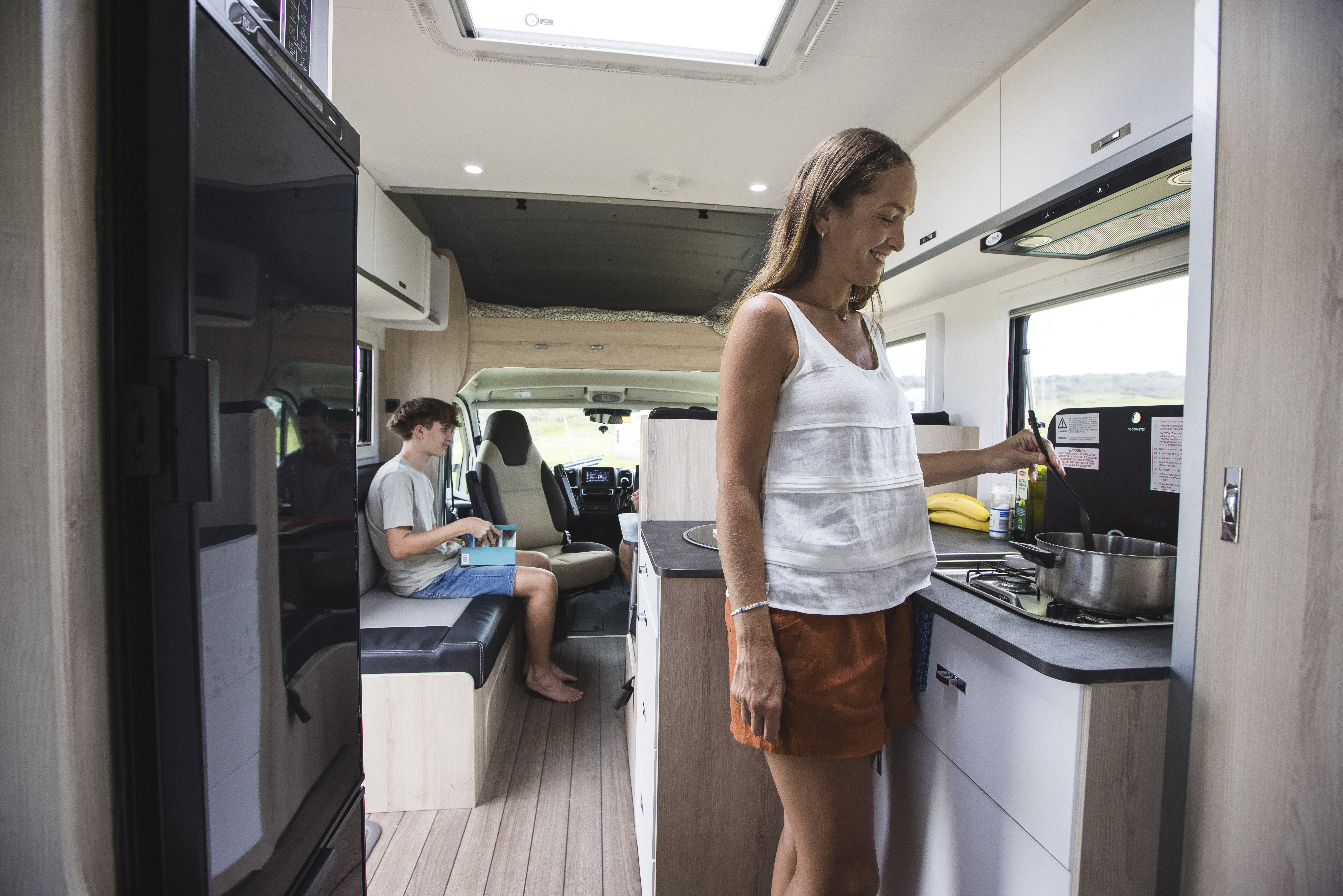 Woman cooking inside a modern campervan kitchen, with another person sitting in the living area reading.