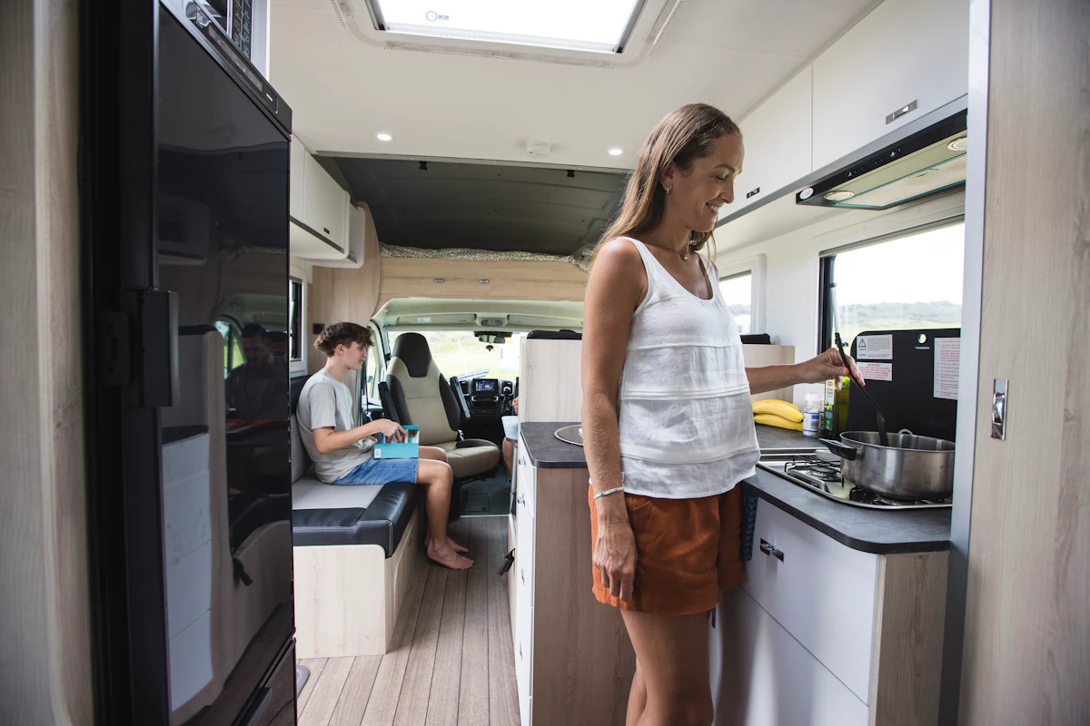 Woman cooking inside a modern campervan kitchen, with another person sitting in the living area reading.