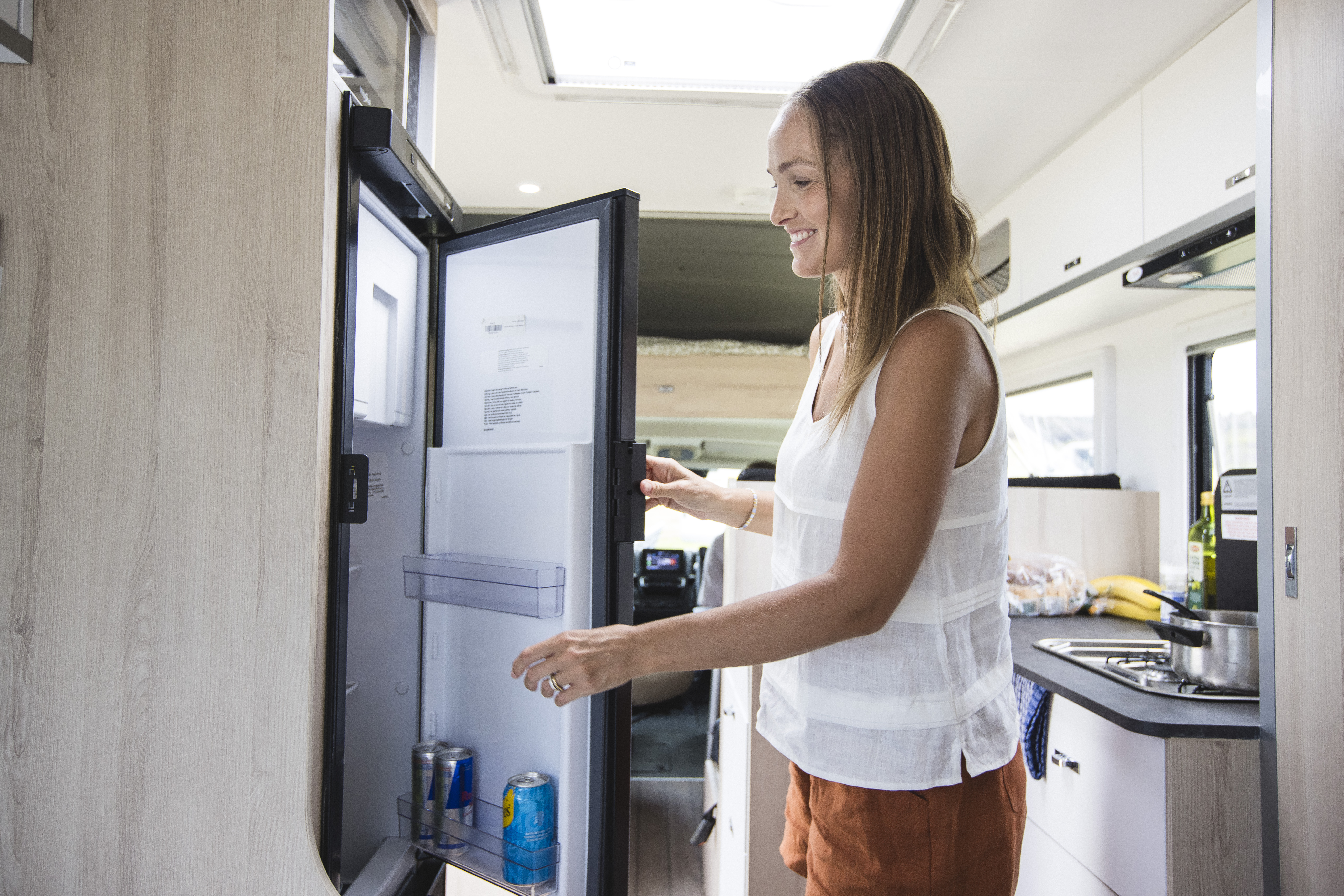 Woman opening a fridge inside a modern campervan kitchen, with fresh fruits and groceries visible on the counter.