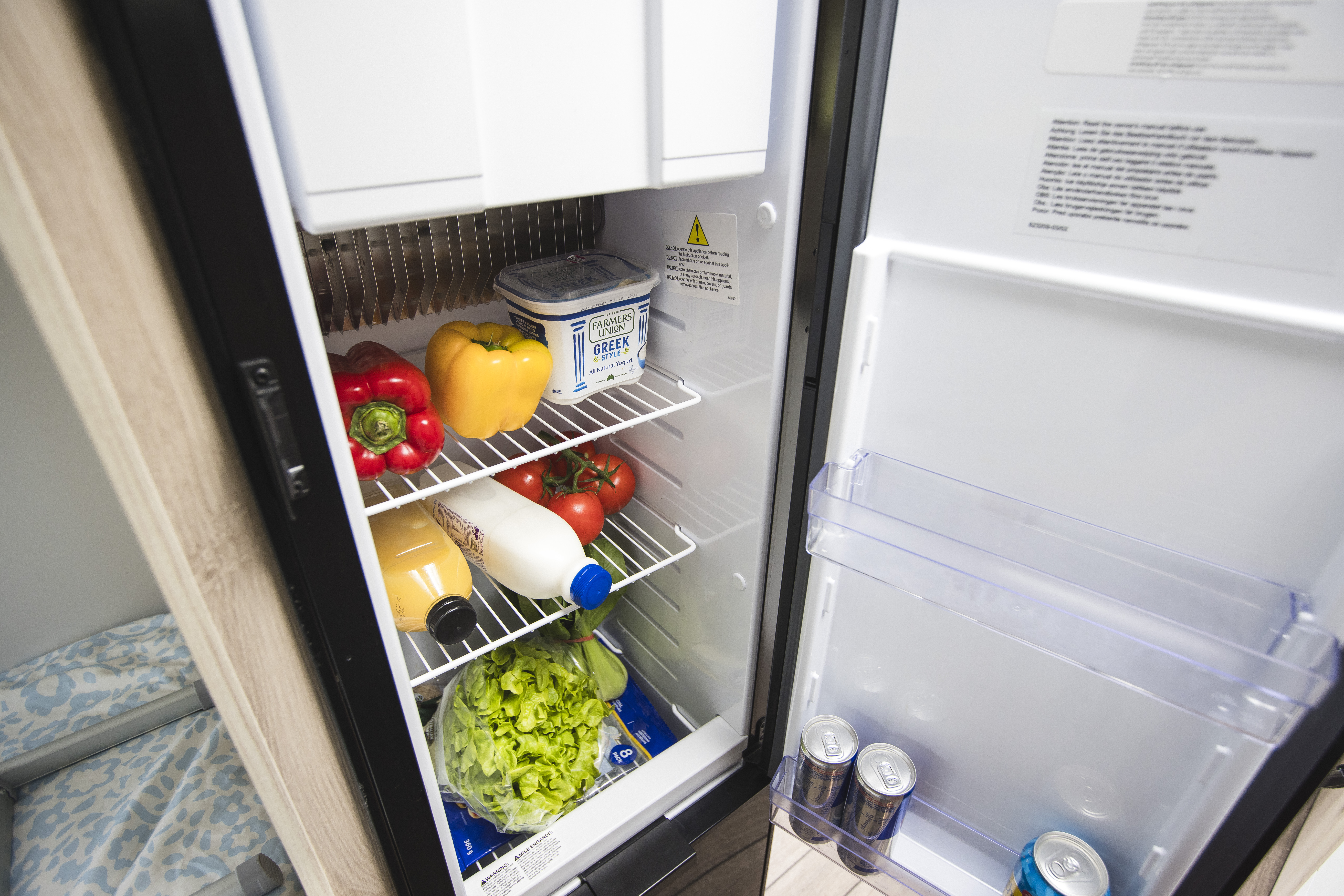 Open campervan fridge containing vegetables, milk, juice, butter, and cans, with a bedroom visible on the left.