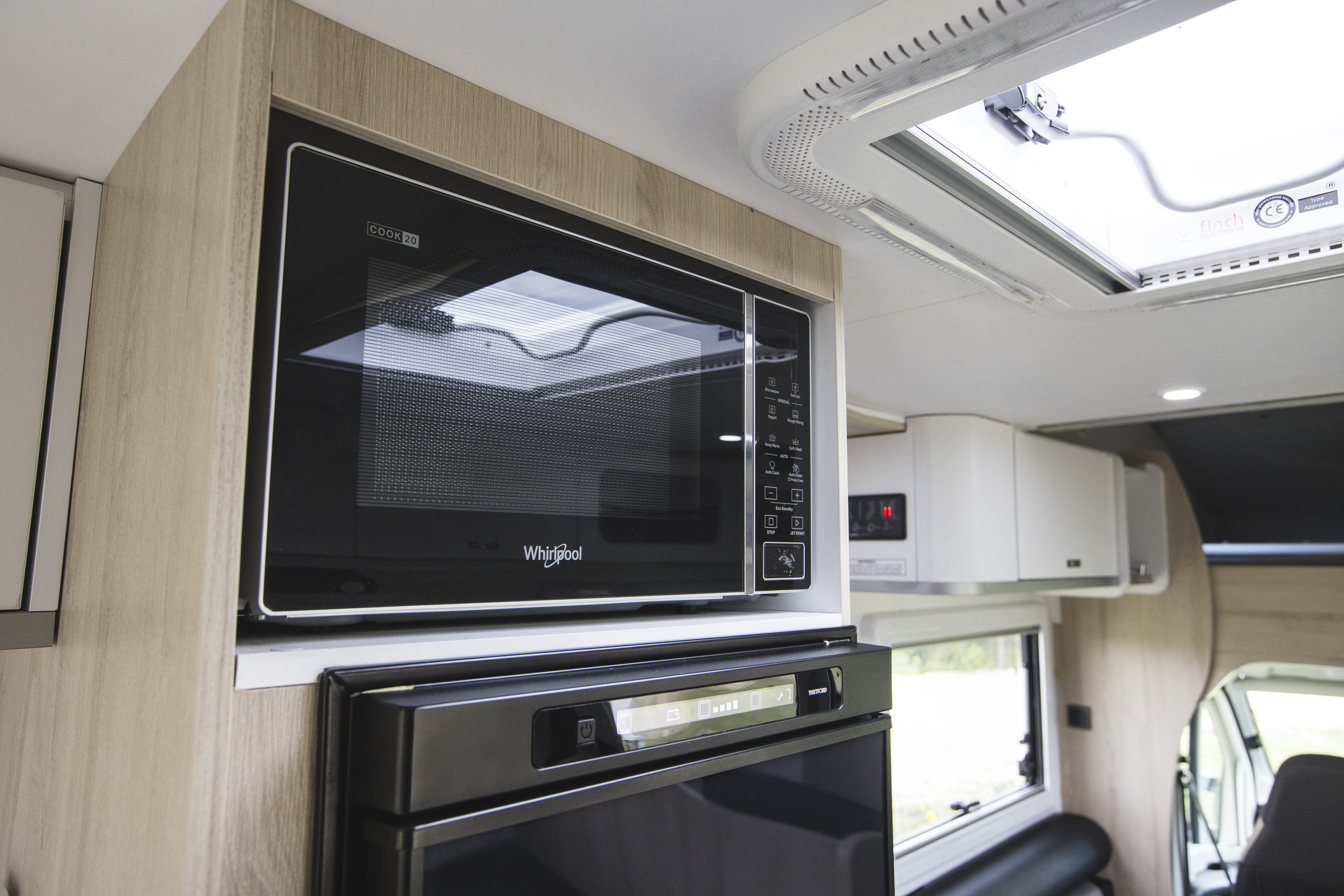 Modern campervan kitchen featuring a built-in Whirlpool microwave above a refrigerator, with natural light from a ceiling window.