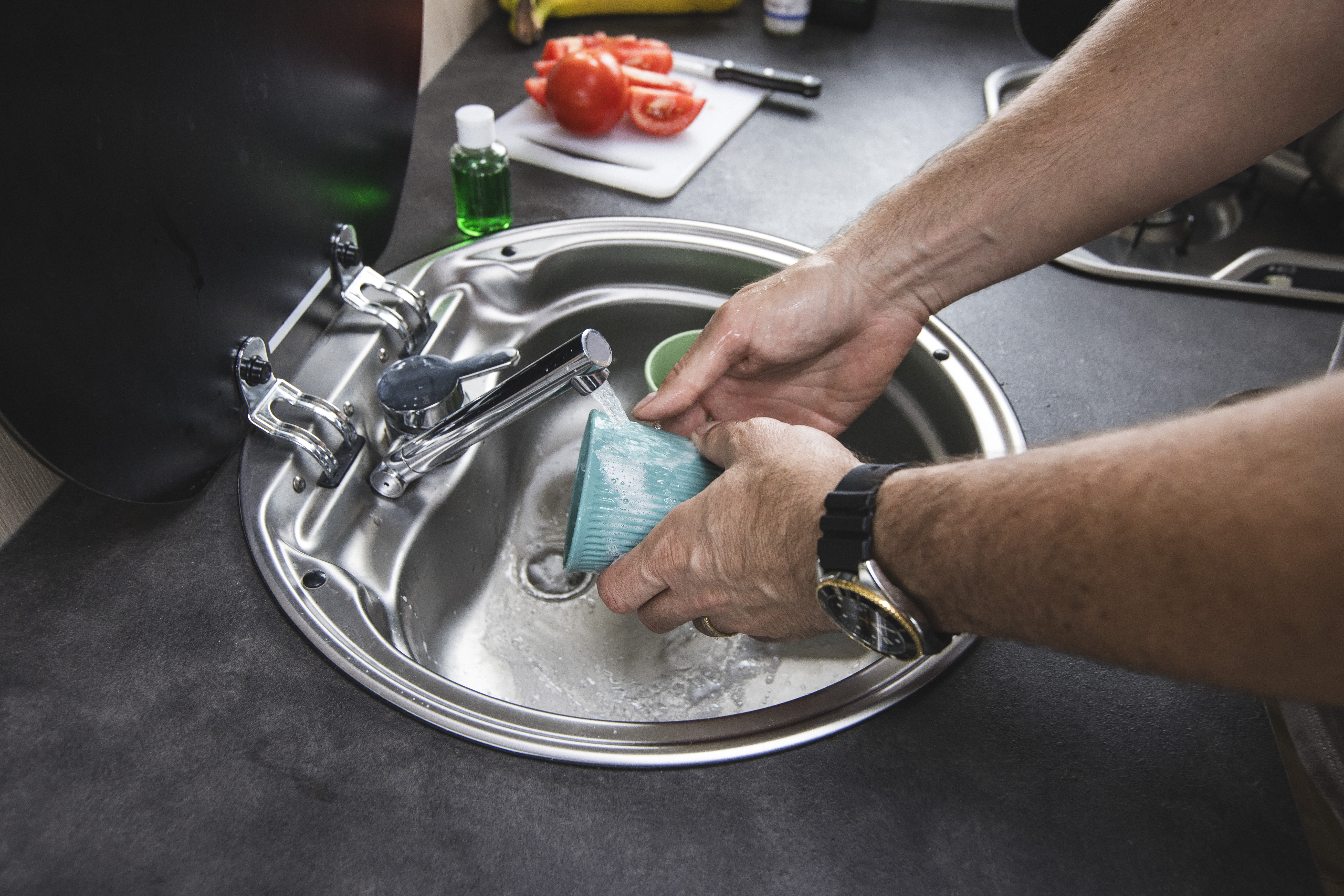 Person washing a cup in a campervan sink with vegetables and dish soap visible in the background.
