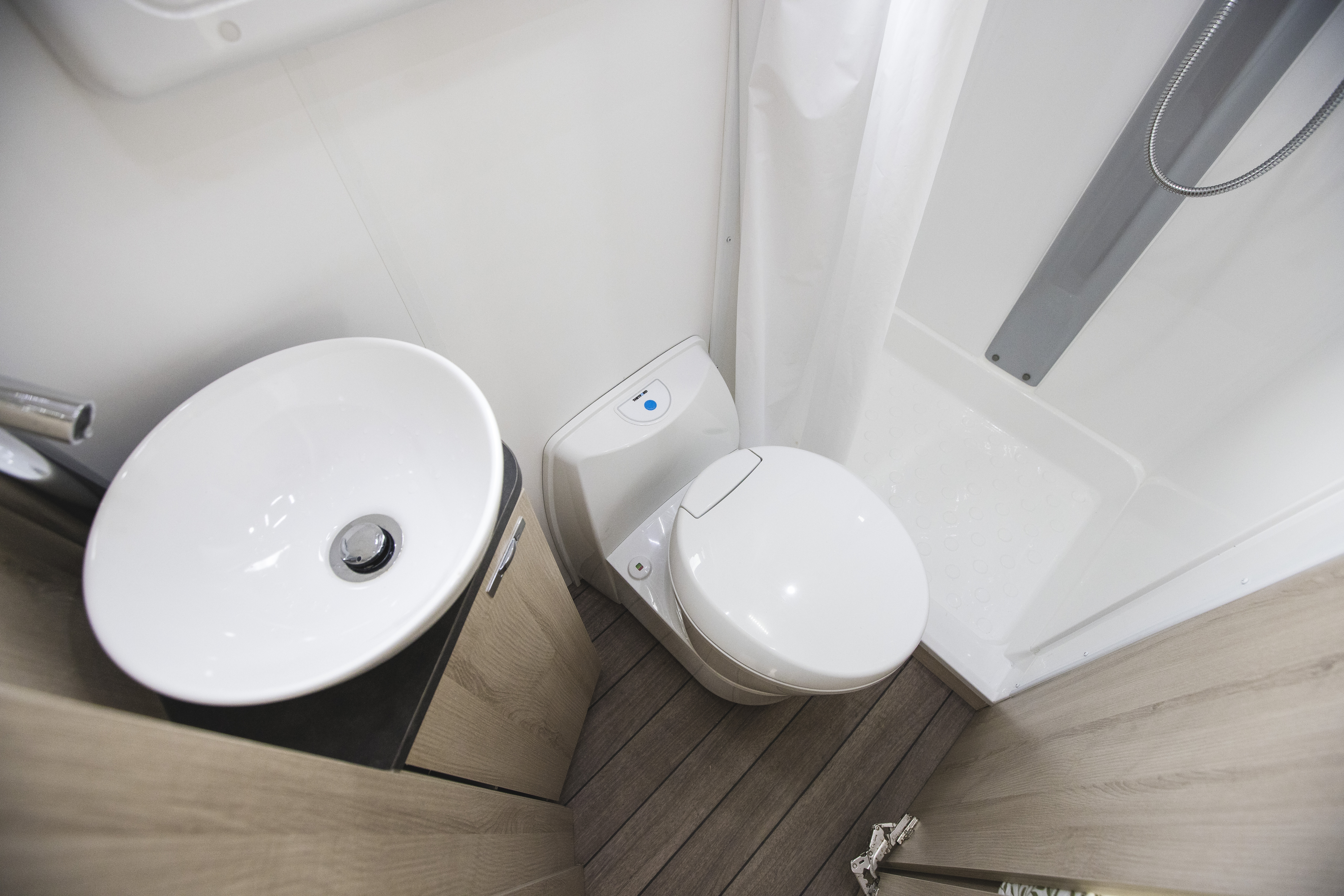 Compact campervan bathroom with a white sink, toilet, and shower area, featuring modern fixtures and wood-style flooring.
