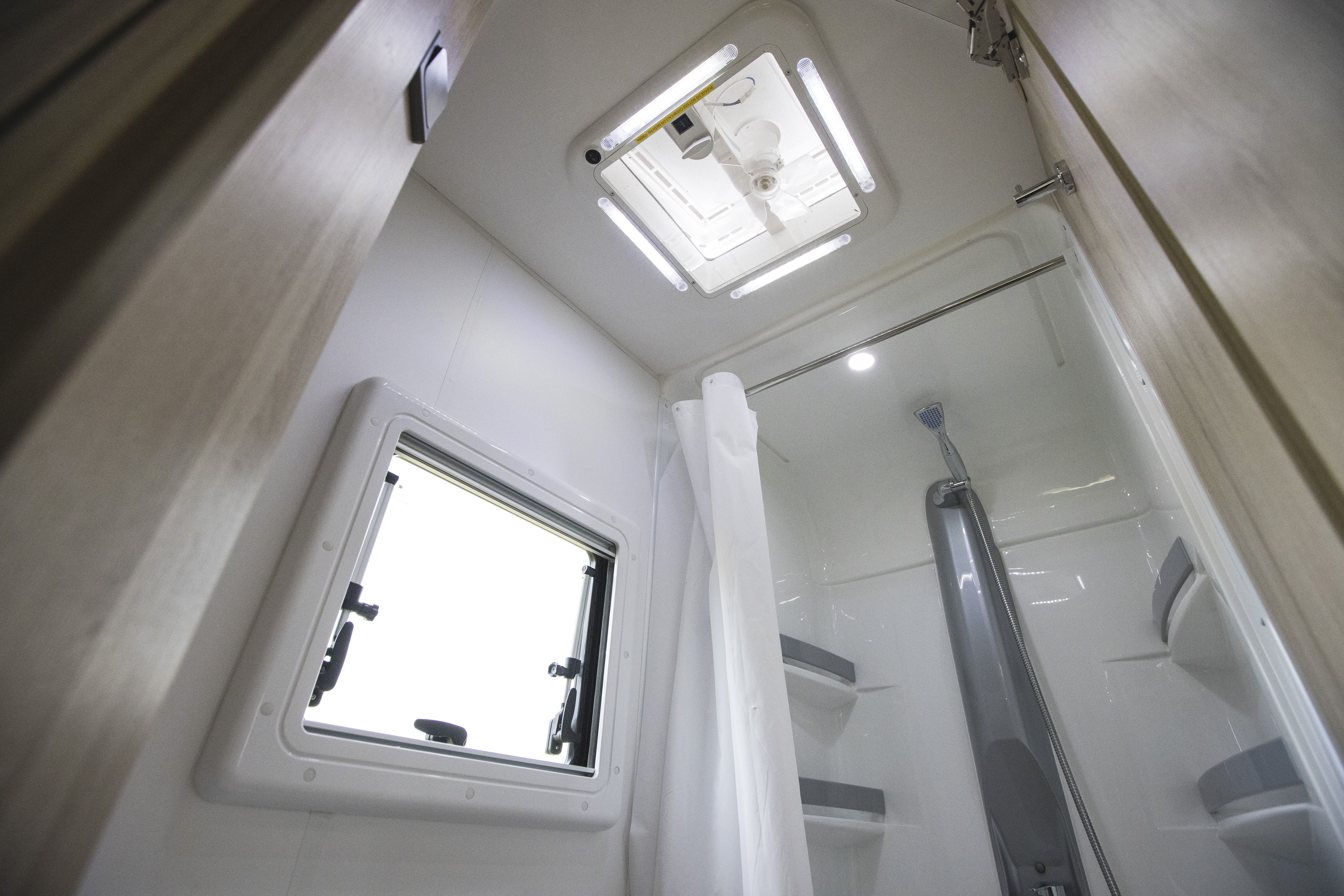 Interior view of a campervan bathroom with shower, ceiling vent, window, and shelves.