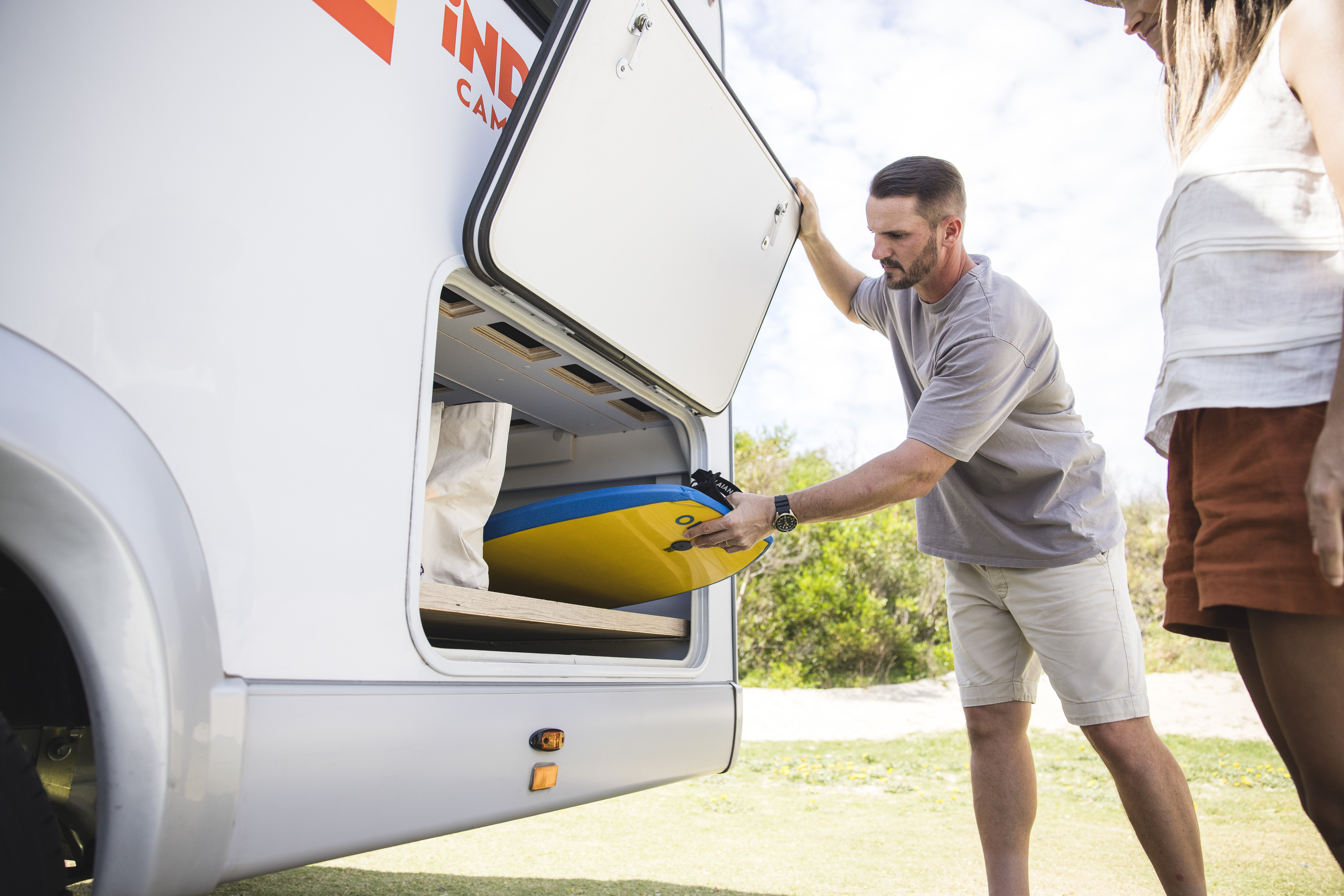 Man loading a surfboard into the rear storage compartment of an Indie Campers motorhome outdoors.