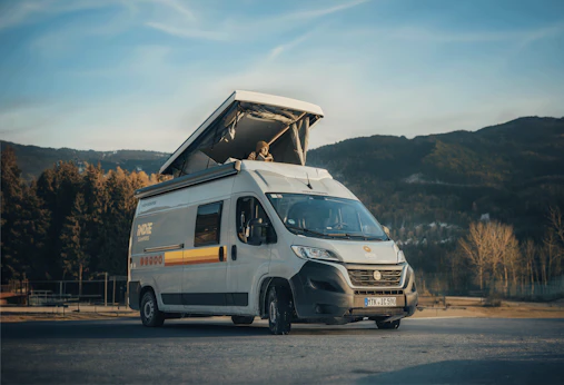 White campervan with raised pop-up roof parked outdoors, surrounded by trees and distant mountains under a blue sky.