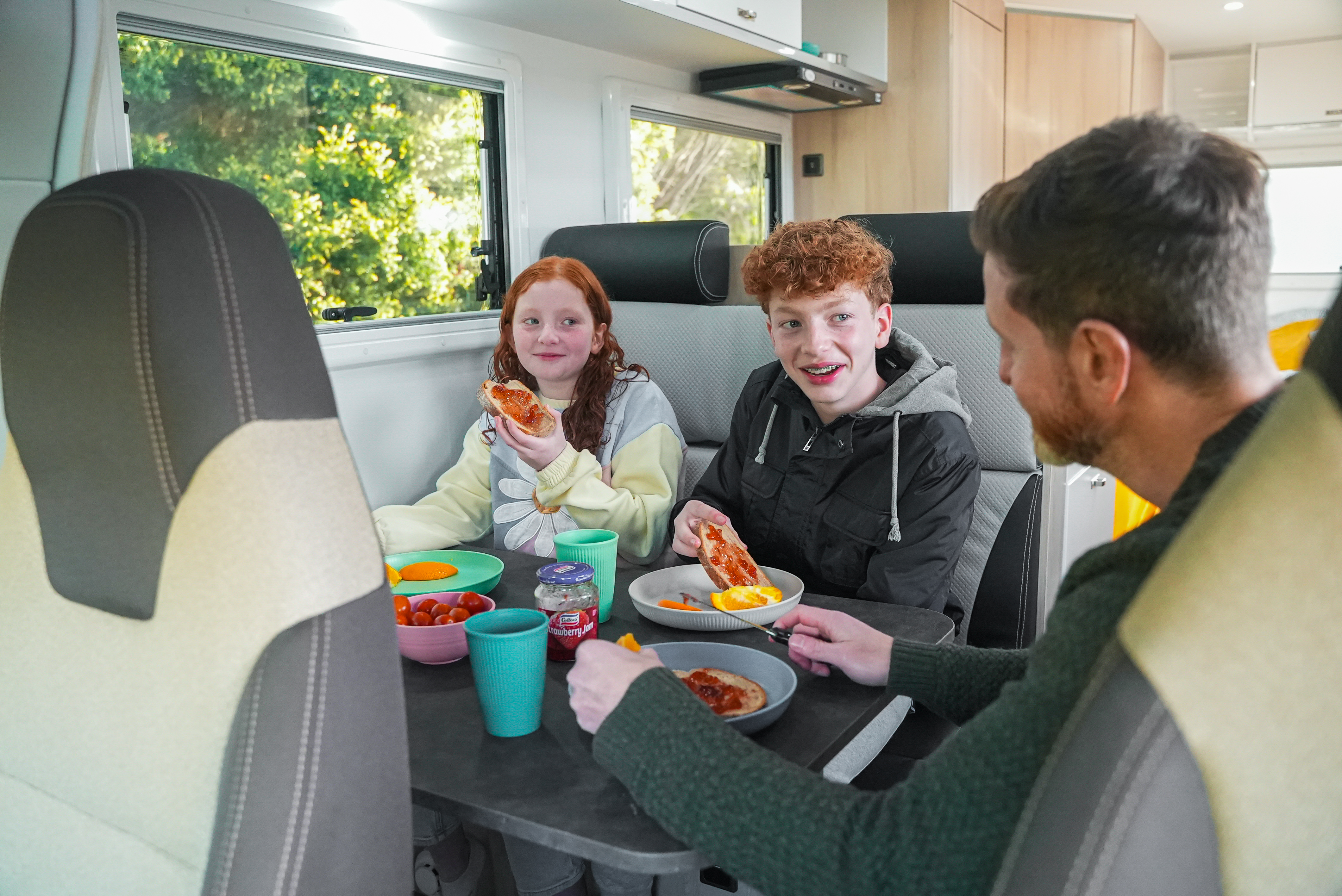 Family enjoying a meal together inside a modern campervan, sitting at the dining table with food and drinks.
