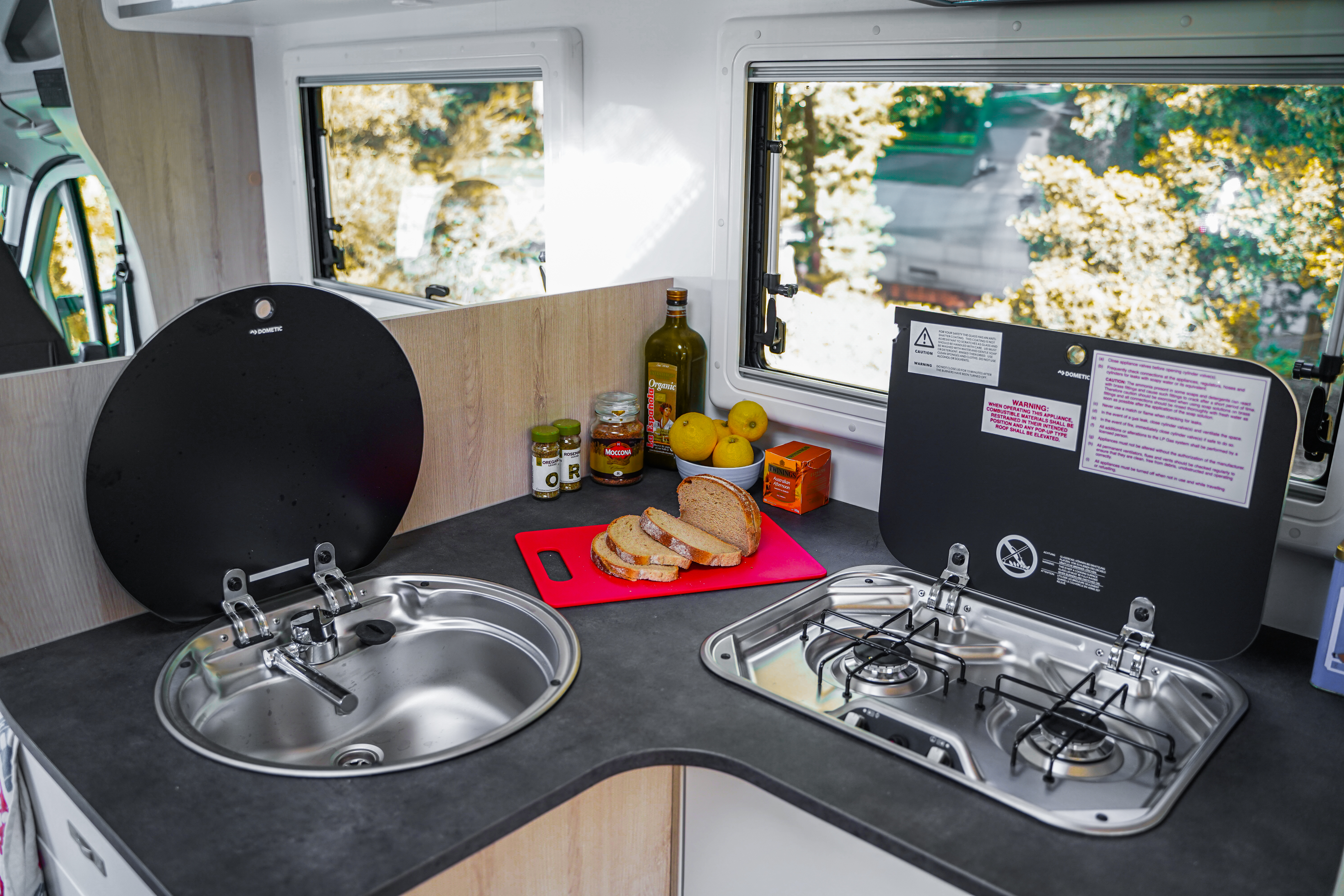 Campervan kitchen area with sink, gas stove, cutting board, bread, fruit, olive oil, and seasonings.