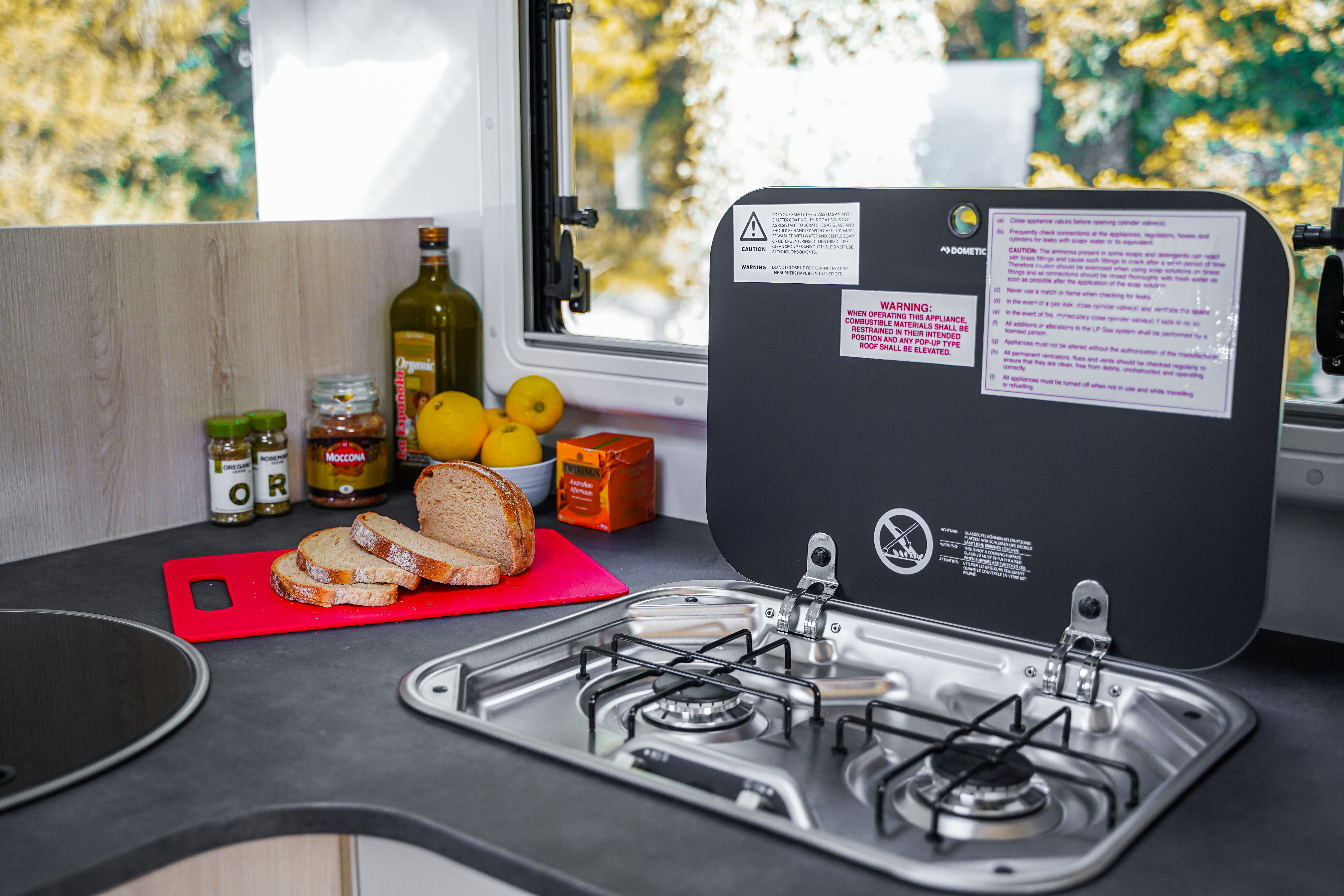 Campervan kitchen with gas stove, bread and ingredients on counter, window view of trees in the background.