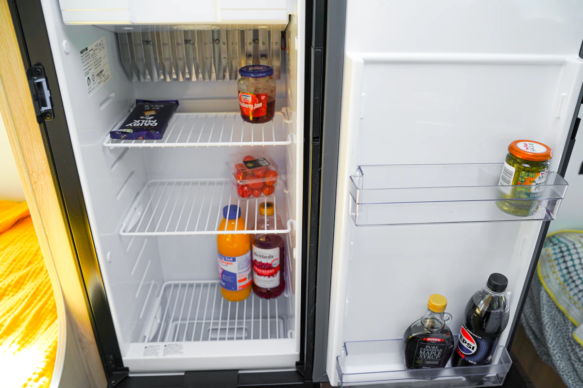 Réfrigérateur de camping-car ouvert avec des boissons, des bocaux et des provisions sur les étagères et la porte, illustrant le rangement typique d’une cuisine de camping-car.