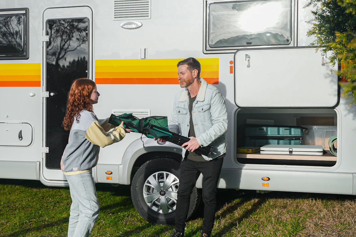 Man and woman unloading camping equipment from a white campervan with yellow and orange stripes, parked on grass.