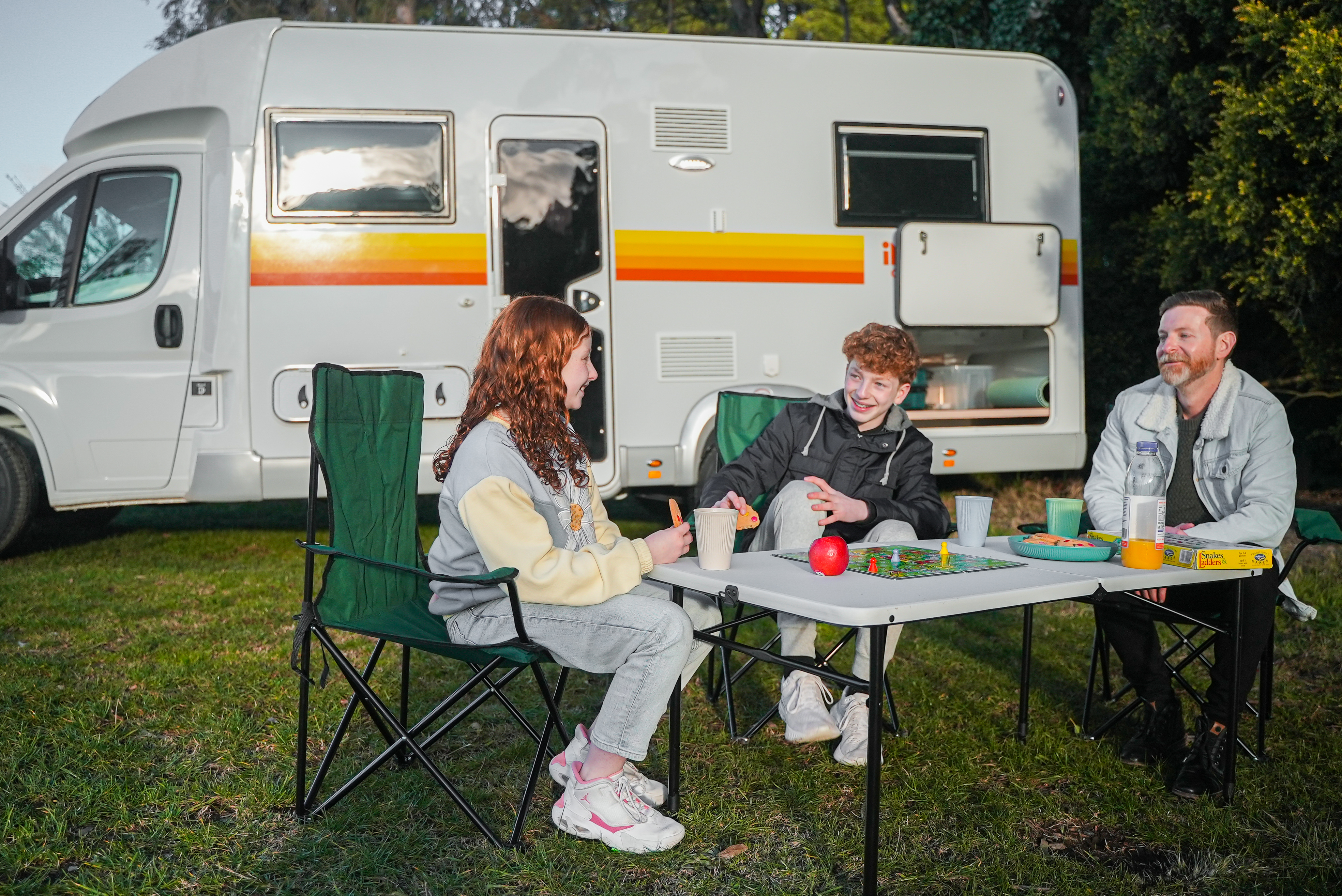 Three people sitting at a table outside a campervan, enjoying drinks and games in a grassy area.