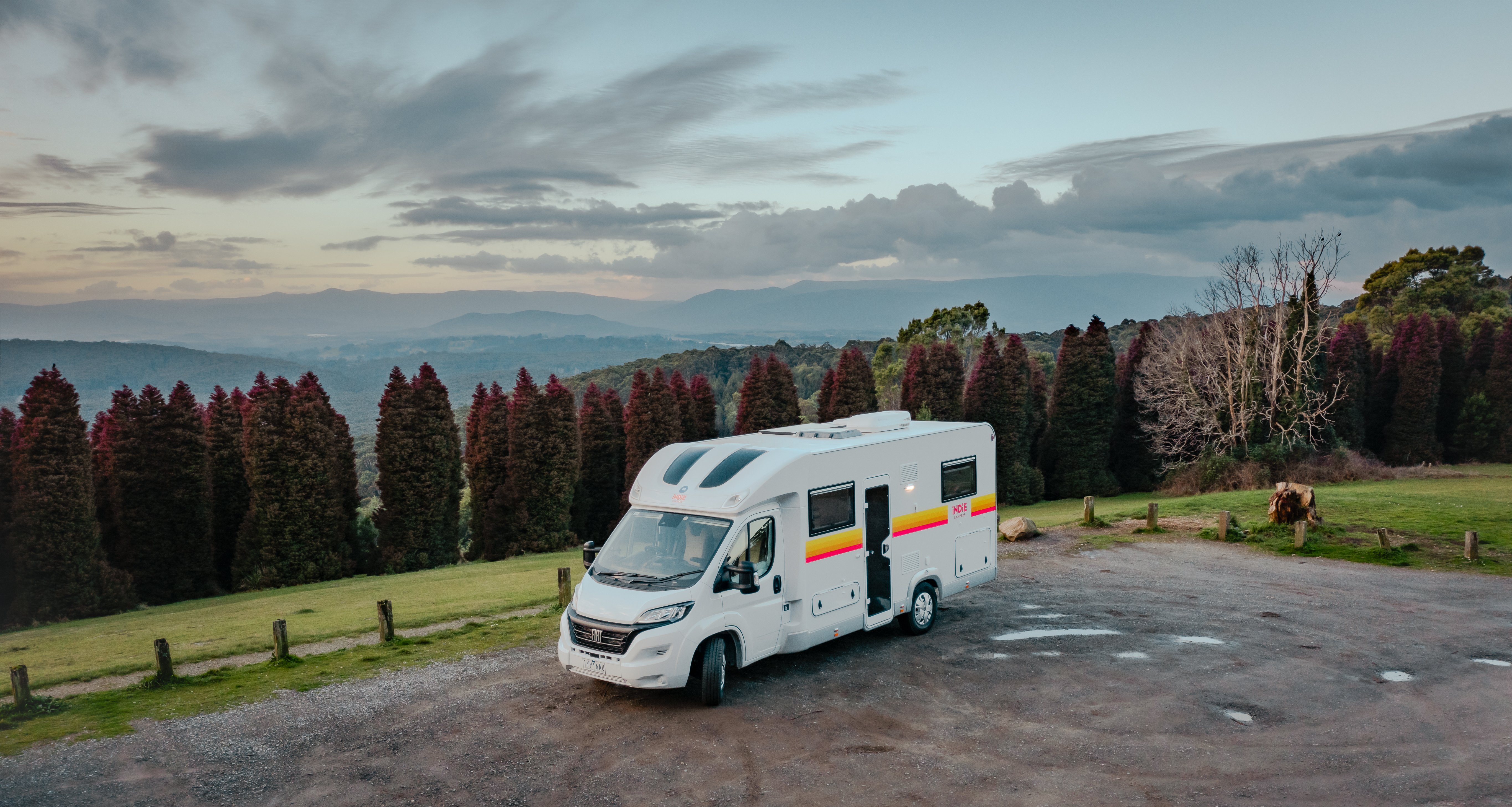 White campervan parked on a scenic overlook with mountains, trees, and cloudy sky in the background.