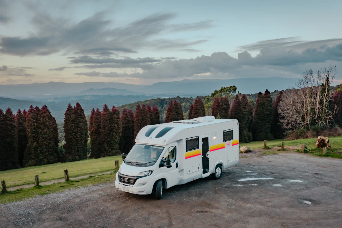 White campervan parked on a scenic overlook with mountains, trees, and cloudy sky in the background.