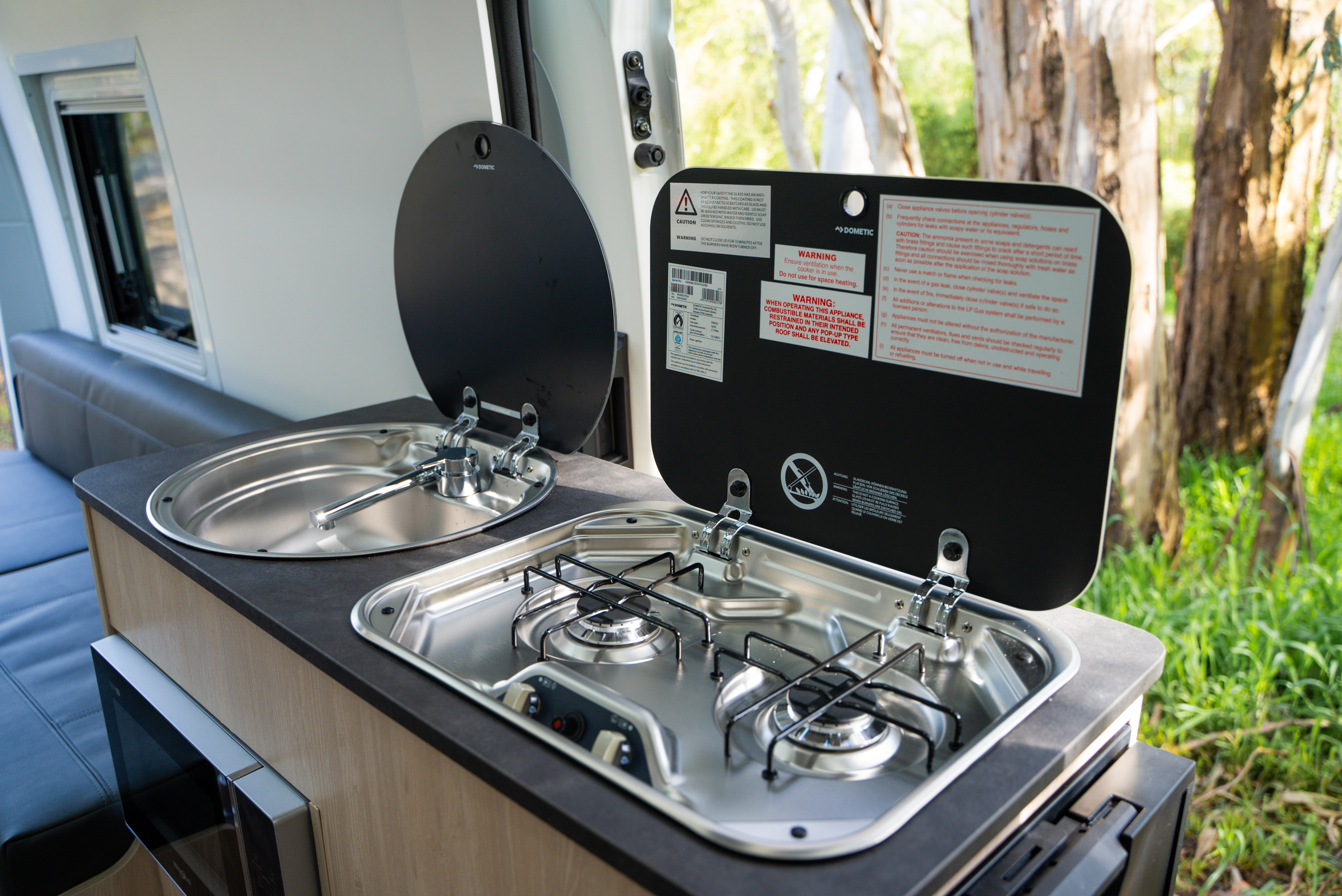 Campervan kitchen with two-burner gas stove and sink, both with open protective covers, surrounded by natural scenery.