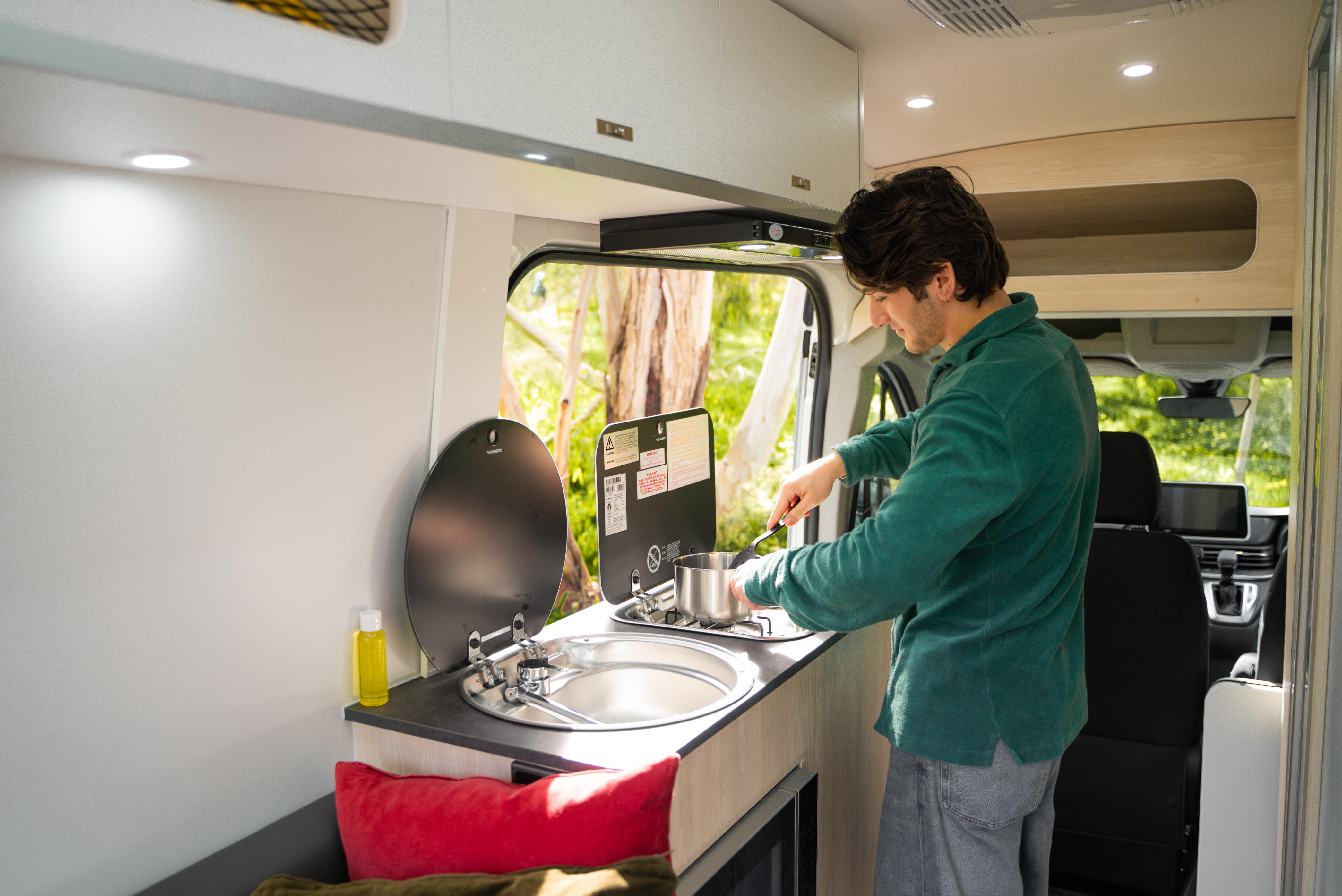 Man cooking at a compact kitchen inside a campervan surrounded by nature, with sunlight coming through the window.