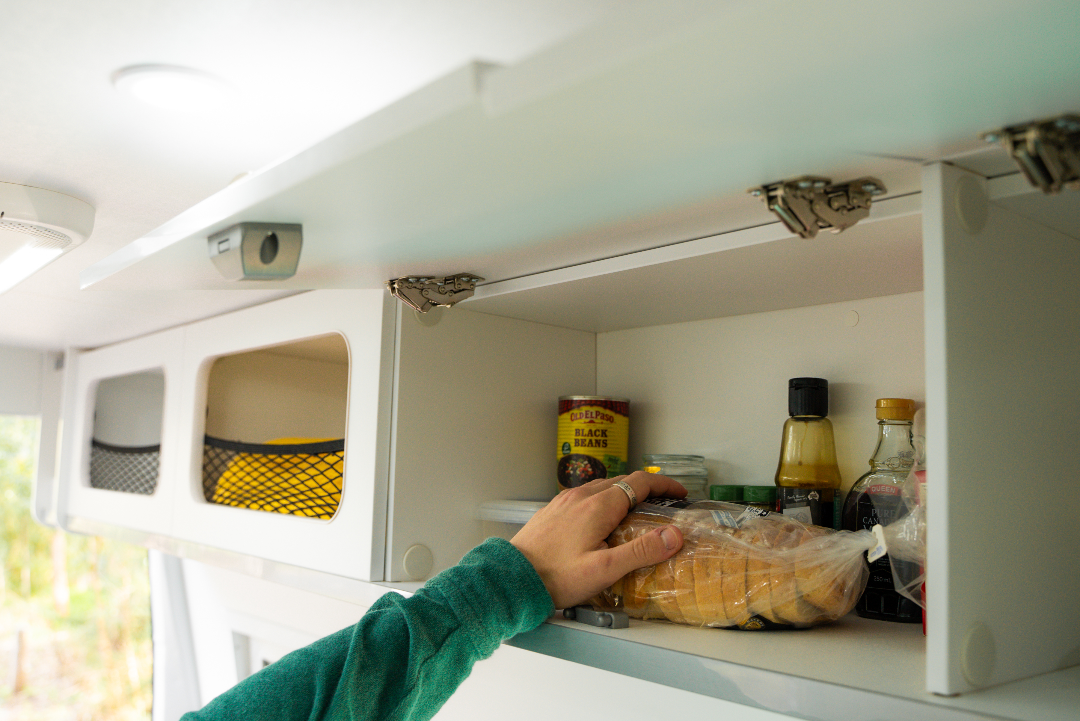 Hand reaching into a campervan kitchen cabinet stocked with food items, groceries, and condiments.