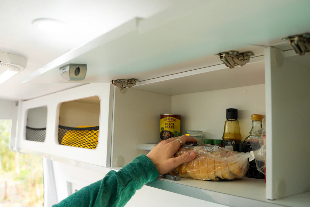 Hand reaching into a campervan kitchen cabinet stocked with food items, groceries, and condiments.