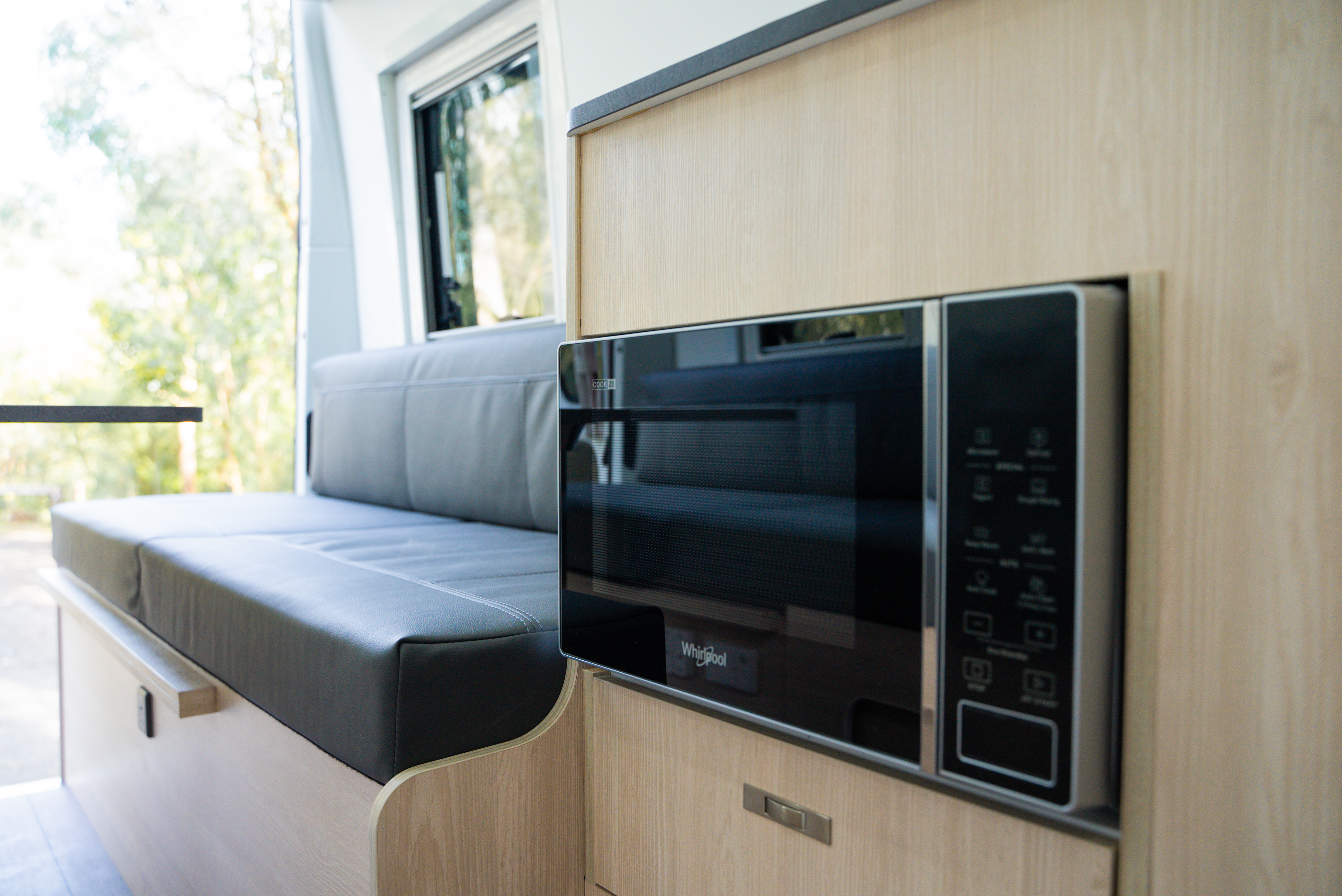 Modern campervan interior featuring a built-in microwave and a cushioned seating area beside a window.