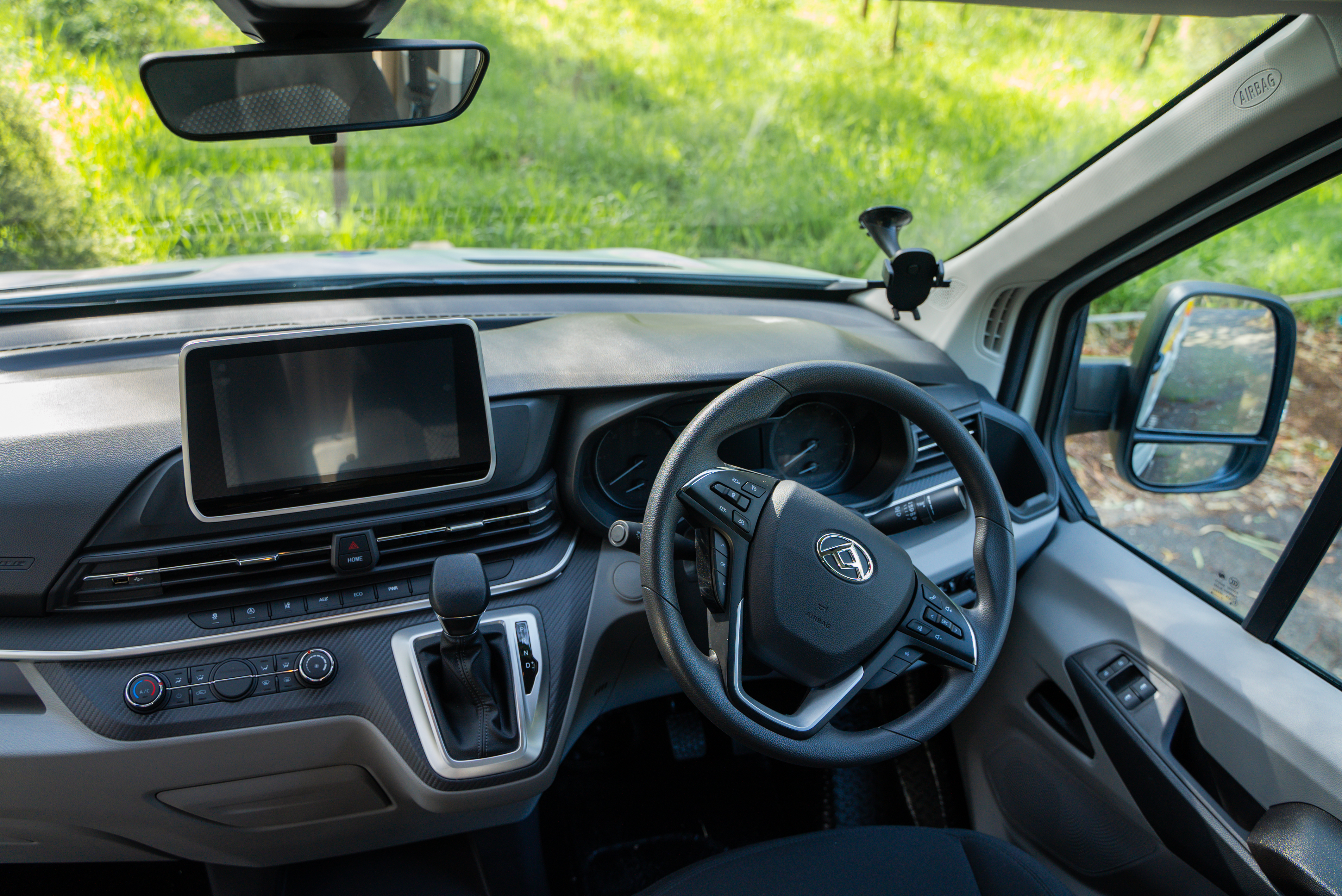 Modern campervan dashboard and steering wheel with touchscreen display, viewed from the driver’s seat, green scenery outside.