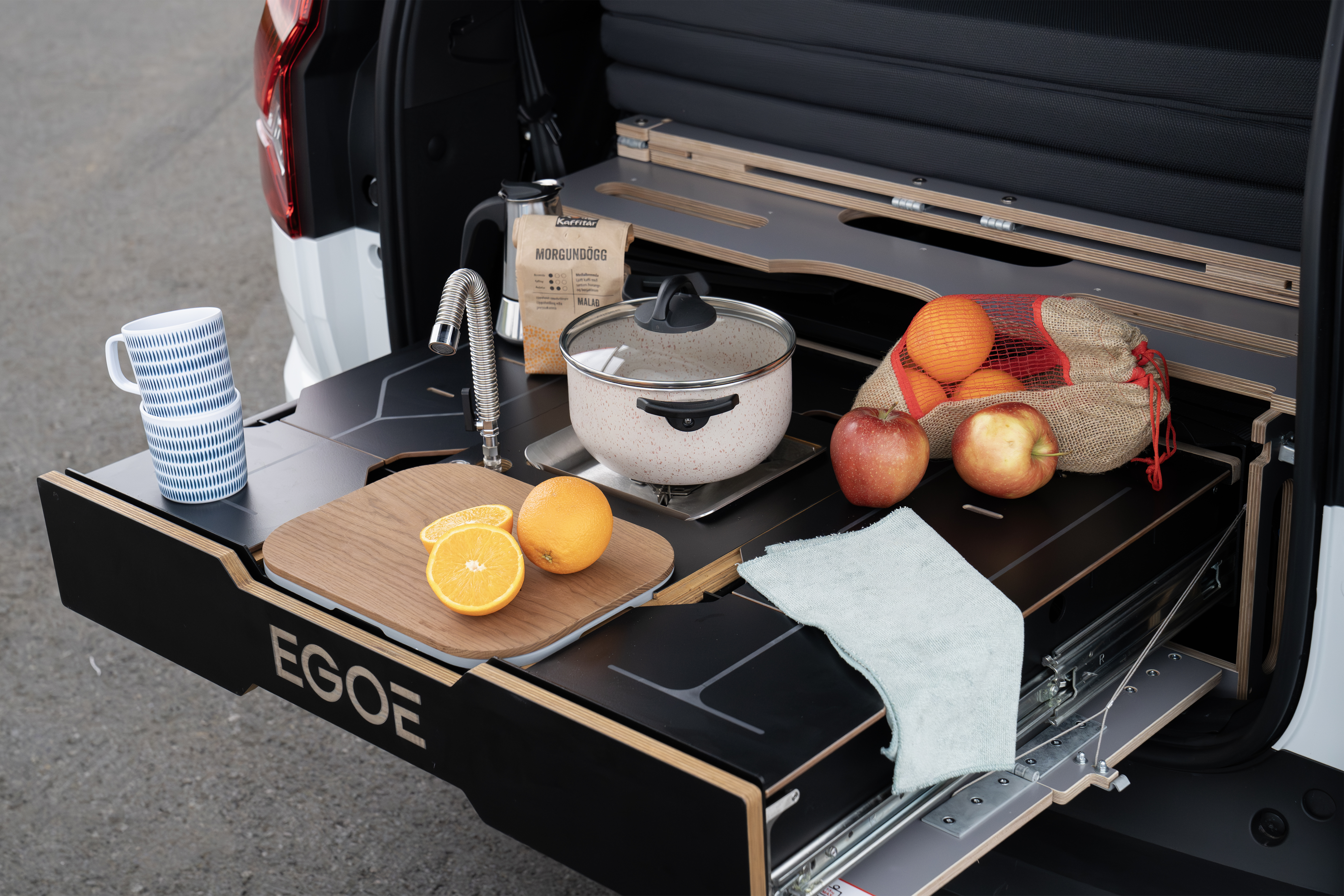 Pull-out camper kitchen with sink, pot, and fresh fruit displayed at the back of a van on a countertop.