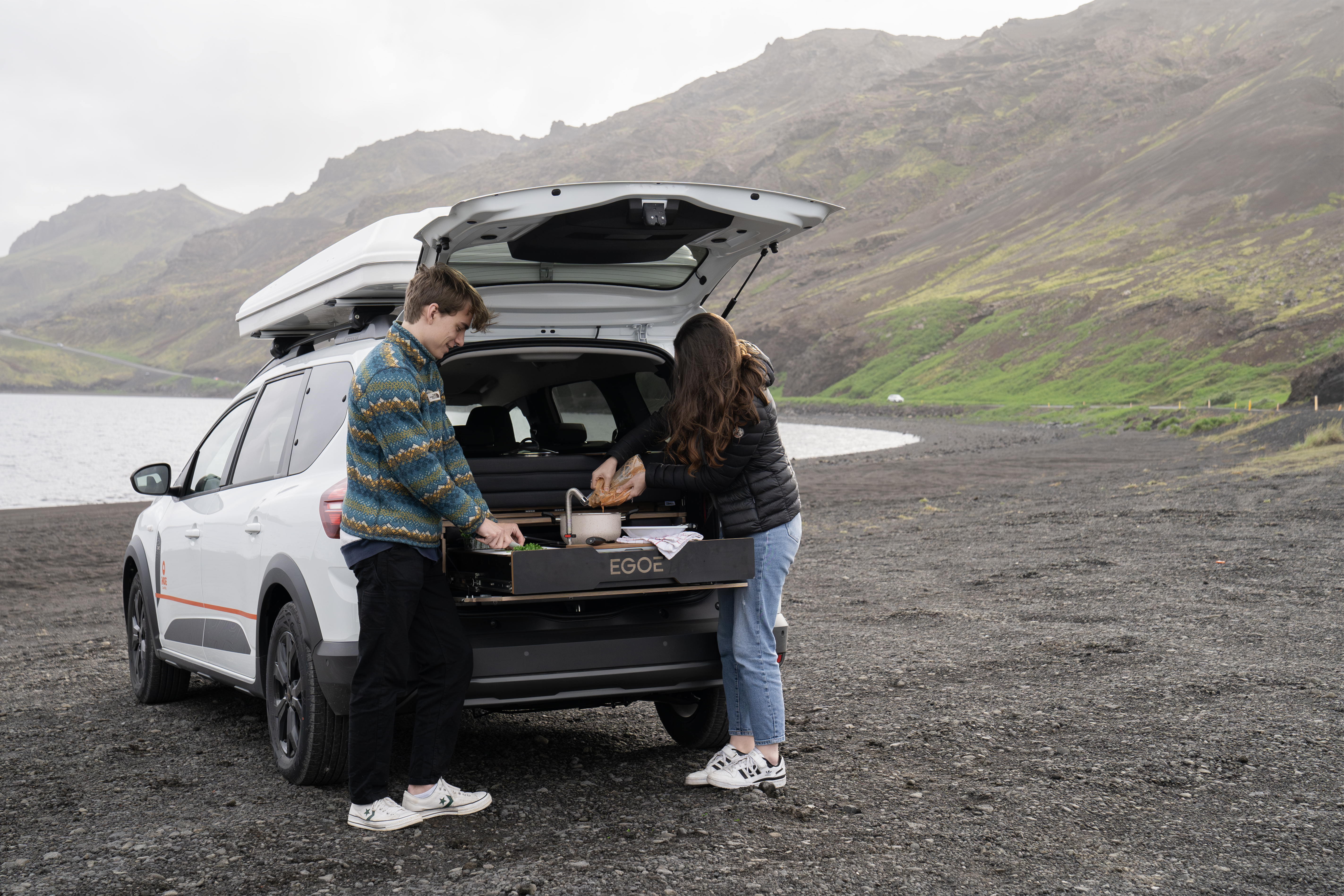 Two people preparing food at the back of a camper van parked on a scenic, rocky shoreline.