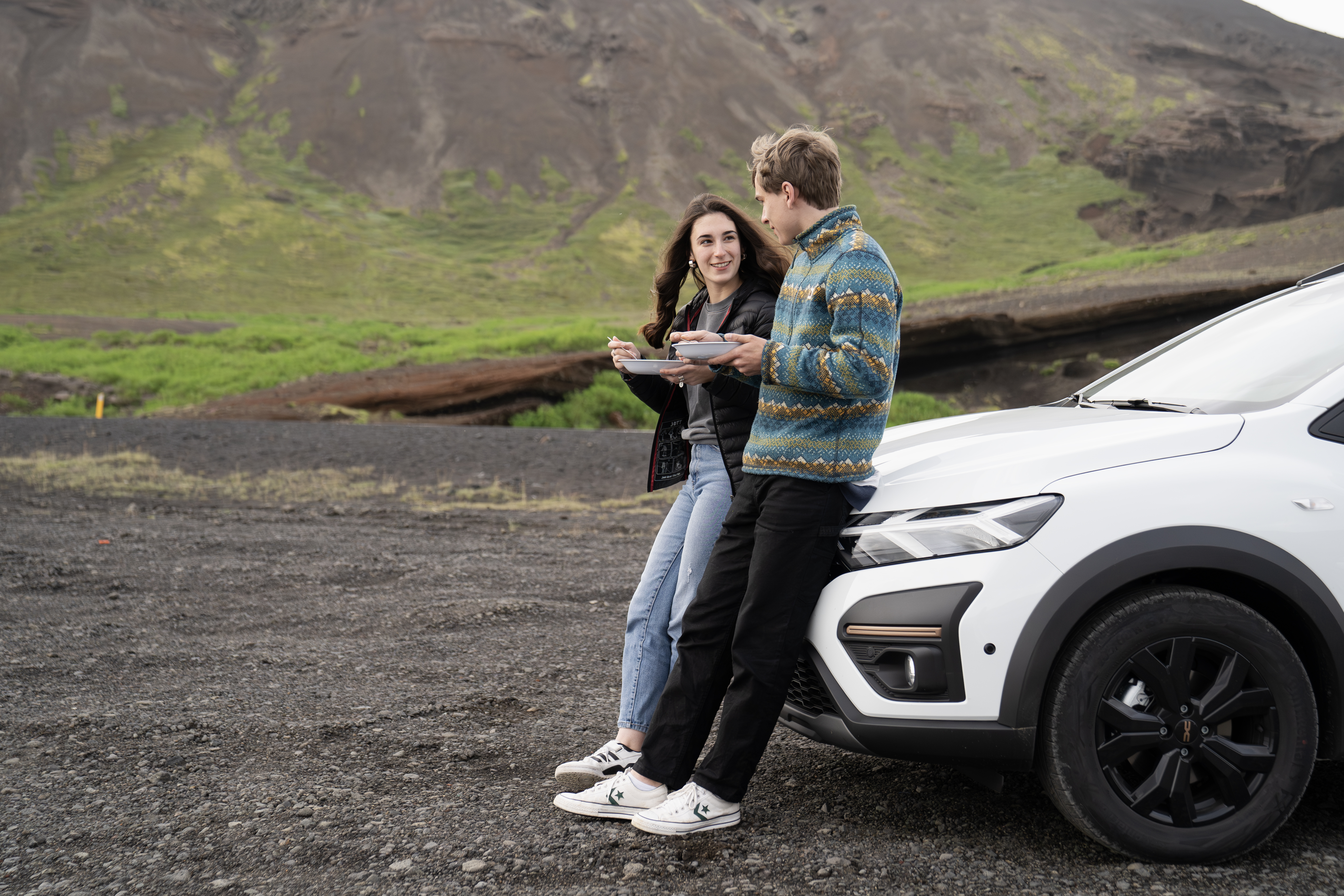 Two people eating by a parked white car with mountains and greenery in the background, outdoors.