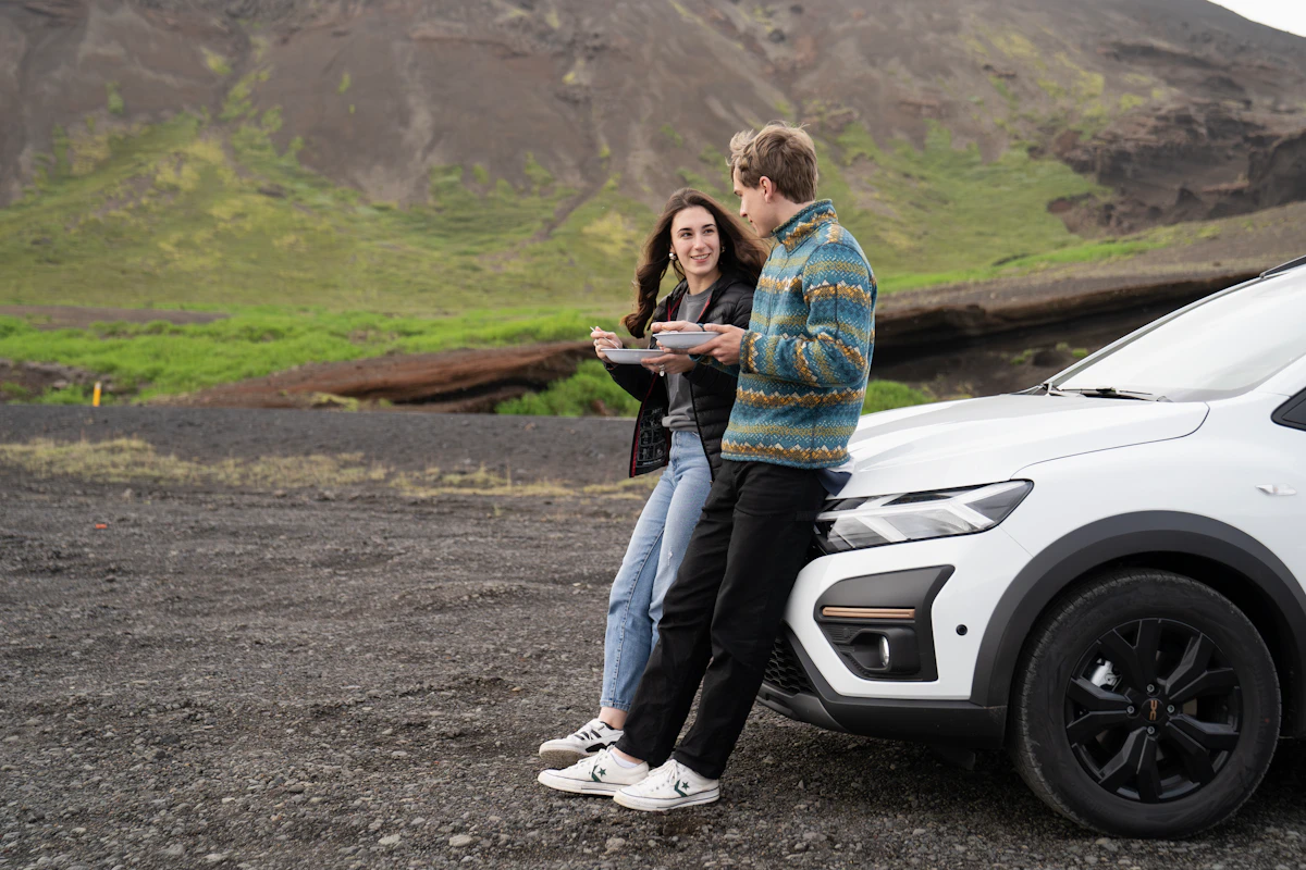 Two people eating by a parked white car with mountains and greenery in the background, outdoors.
