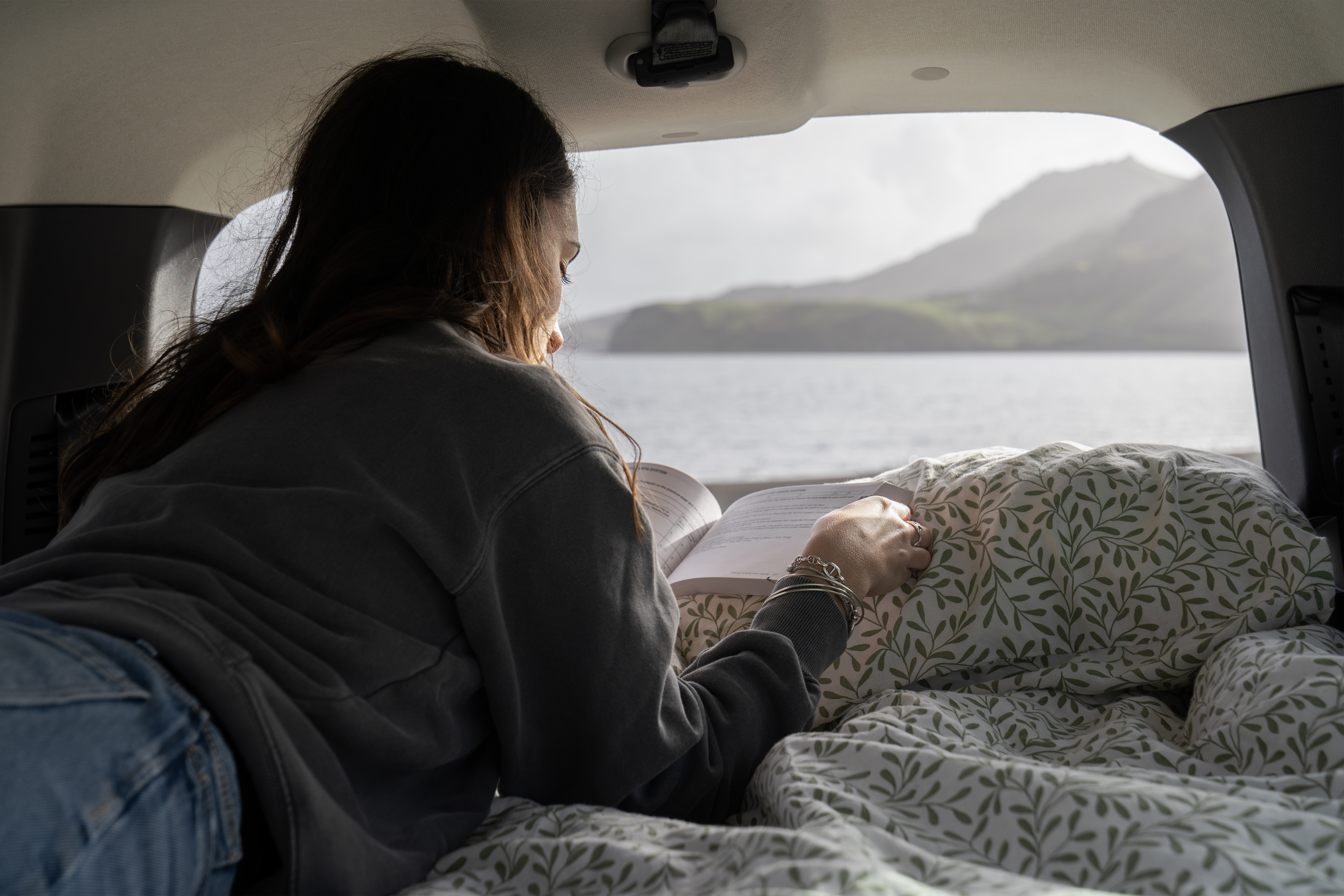 Person reading in campervan bed, overlooking a scenic lake and mountains through the rear window.
