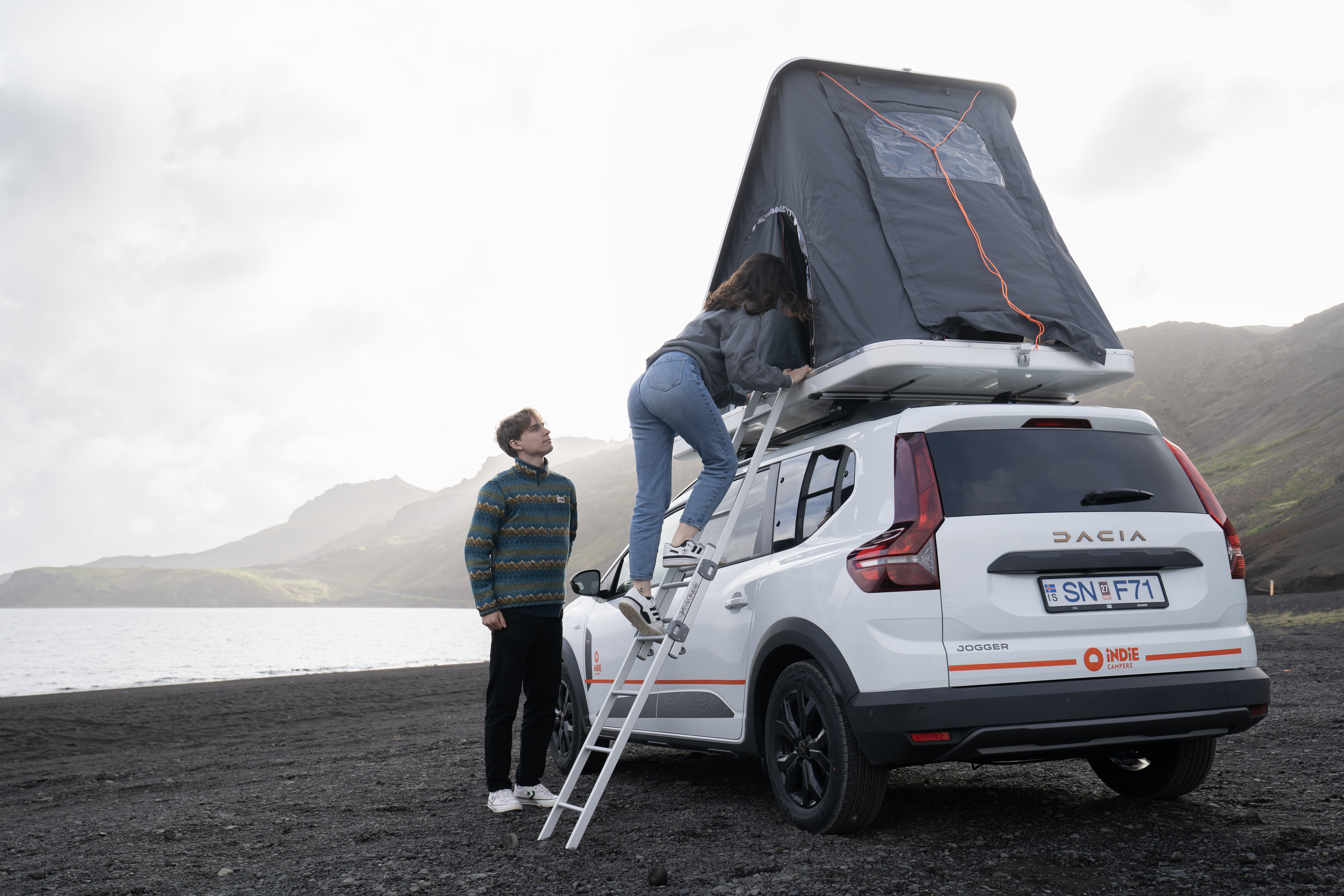 Person climbs rooftop tent on camper van parked by lake, with another person standing beside on black sand beach.