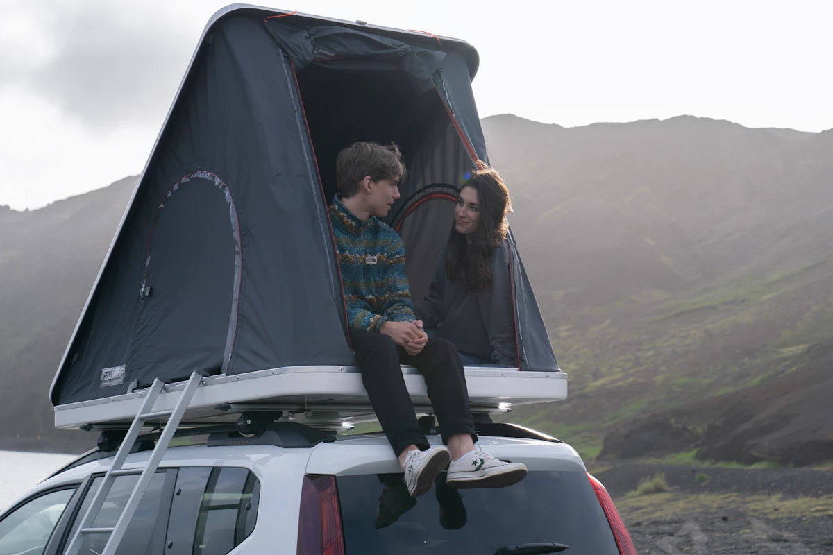 Two people sitting in a rooftop tent on a campervan, overlooking a scenic mountain landscape at dusk.
