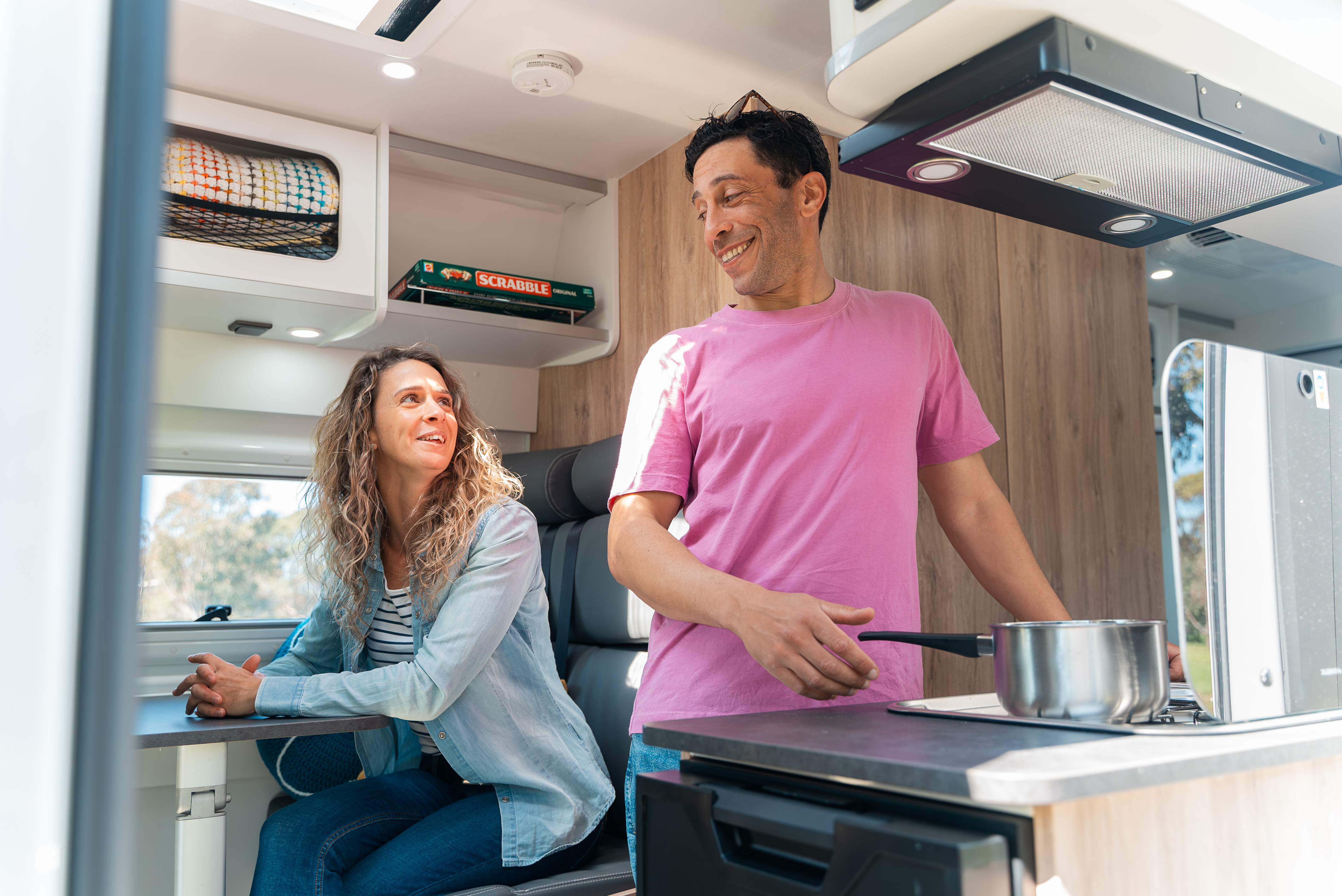 Two people smiling inside a modern campervan kitchen, with one person cooking and the other sitting at a table.