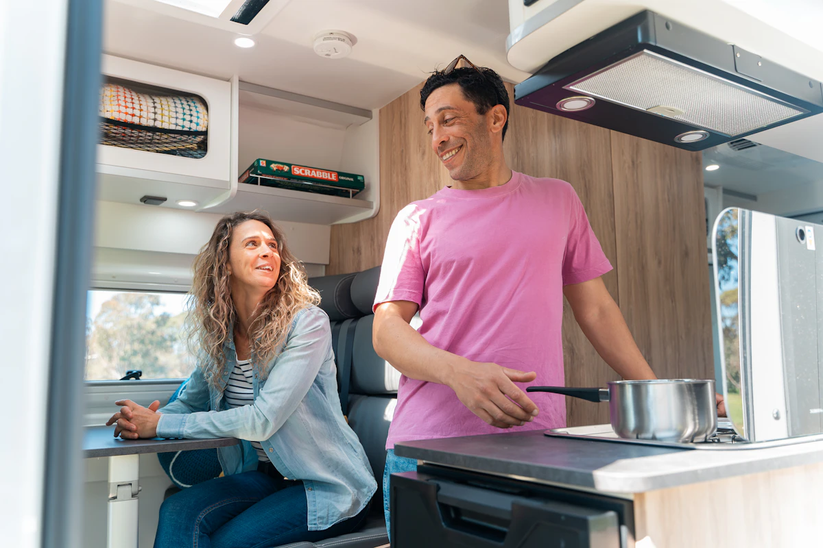 Two people smiling inside a modern camper van kitchen, with one person cooking and the other sitting at a table.