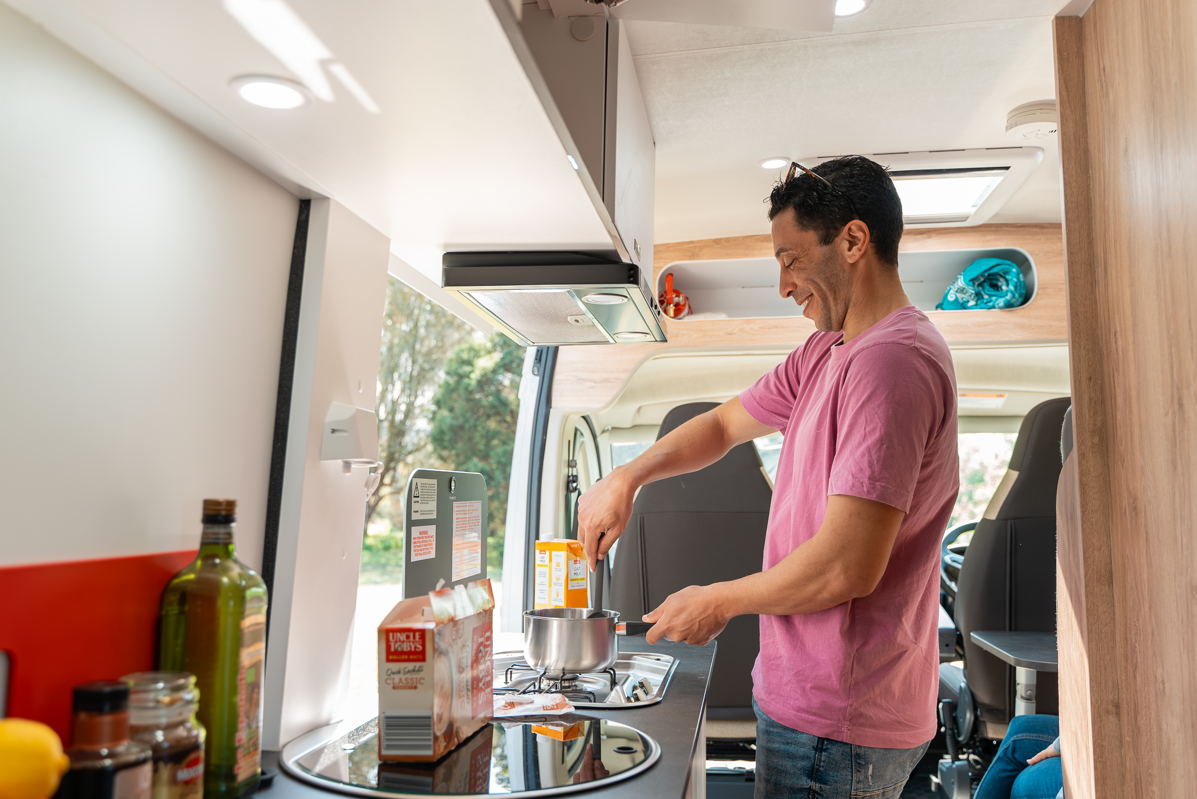 Person cooking in a camper van kitchen with various ingredients and utensils on the counter, daylight outside window.
