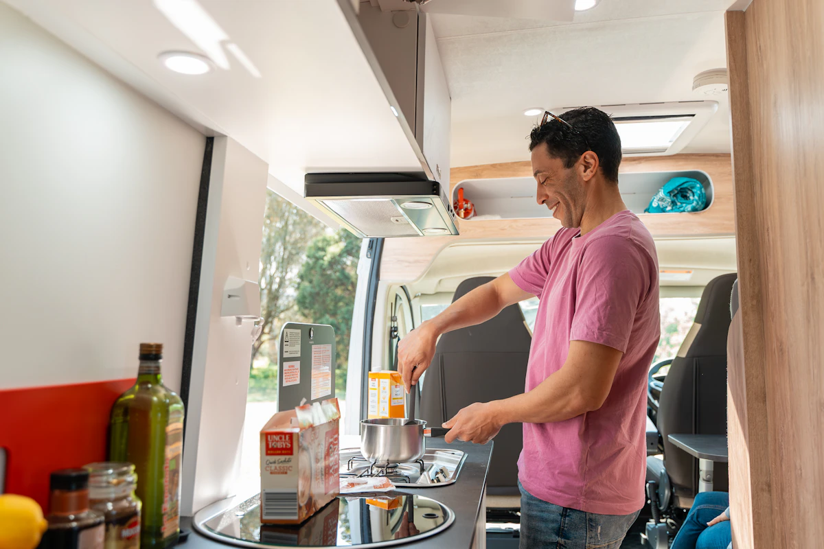 Person cooking in a camper van kitchen with various ingredients and utensils on the counter, daylight outside window.