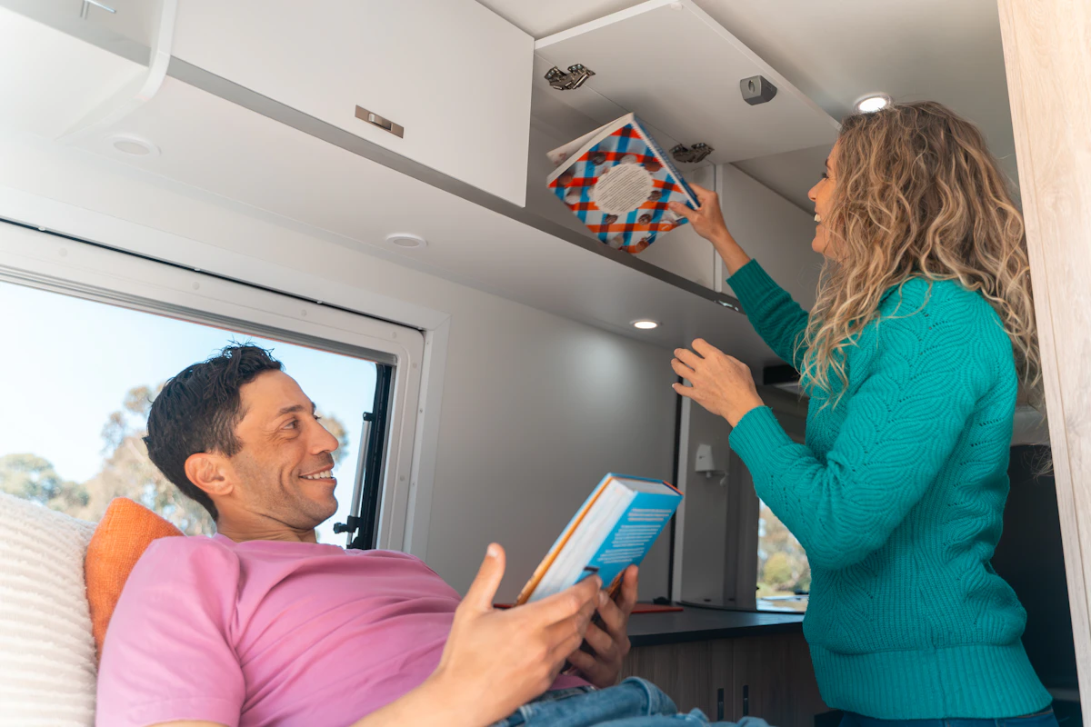 Man relaxing and reading in campervan while woman stores a book in overhead cabinet, bright and cozy interior.