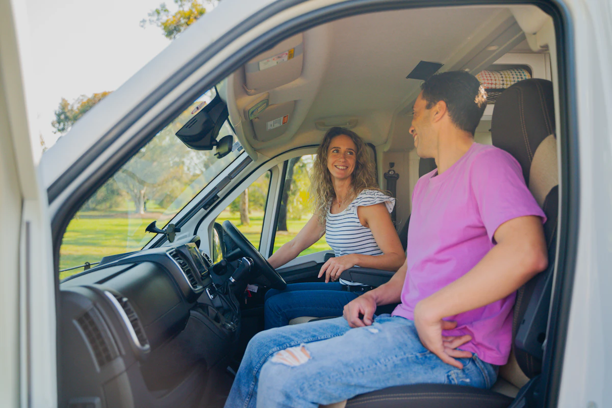 Two people sitting in the front seats of a camper van, smiling and enjoying a sunny day outdoors.