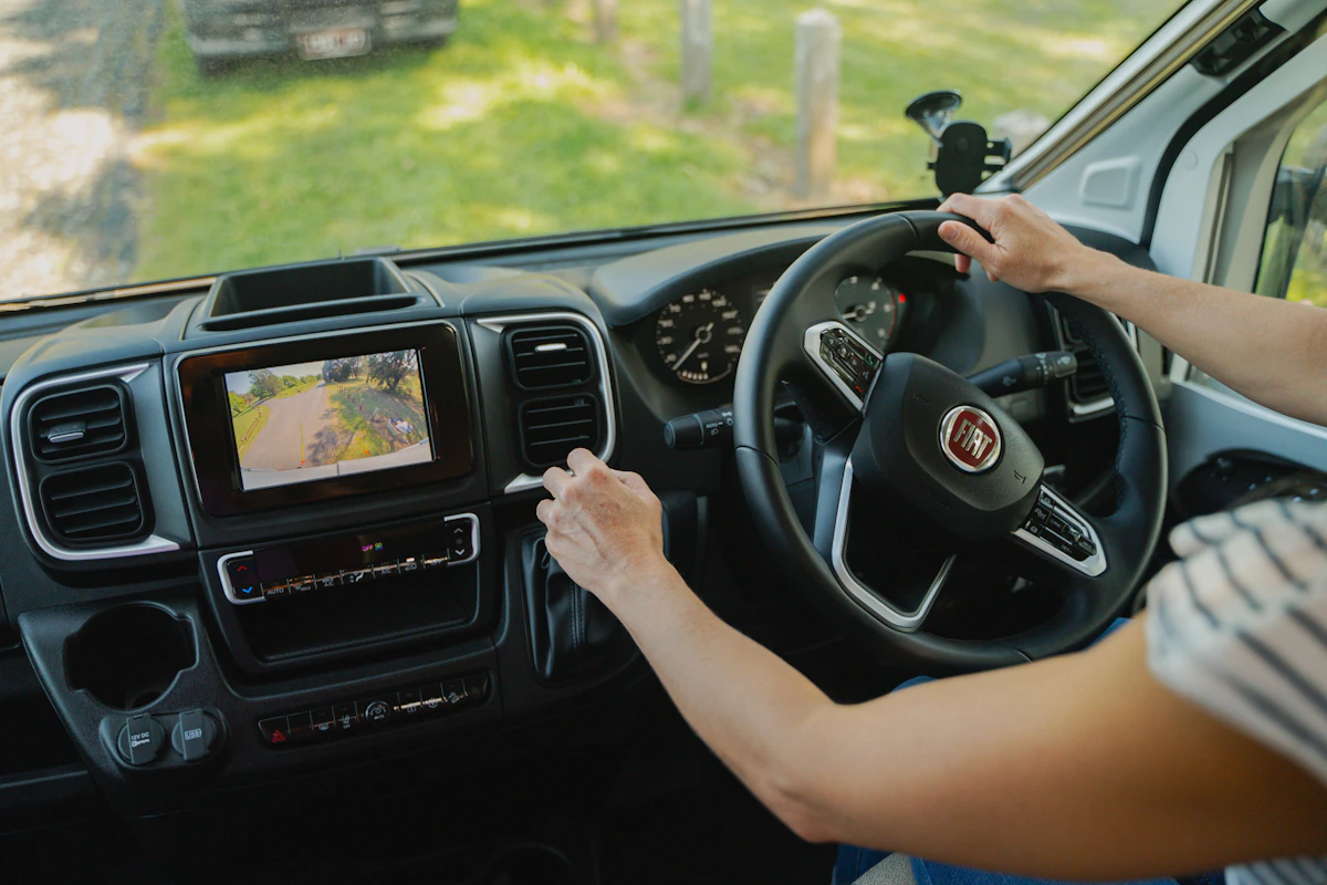 Person driving a Fiat camper van, adjusting the dashboard screen with a rearview camera display.