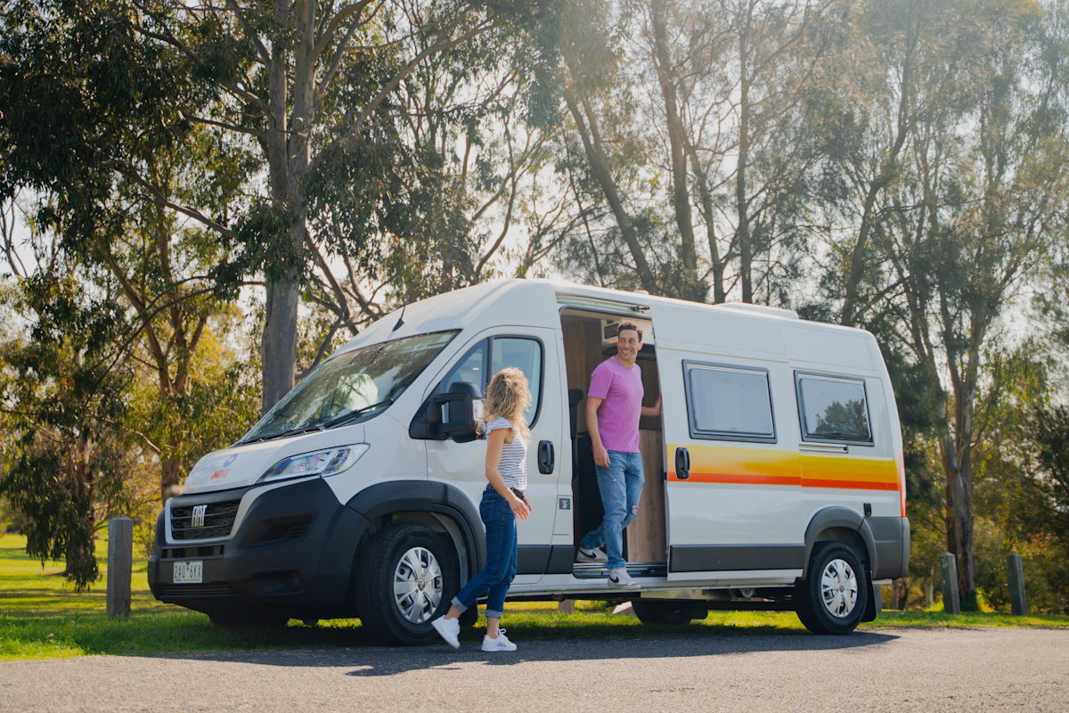 Two people stand by a white campervan parked outdoors with trees and sunlight in the background.